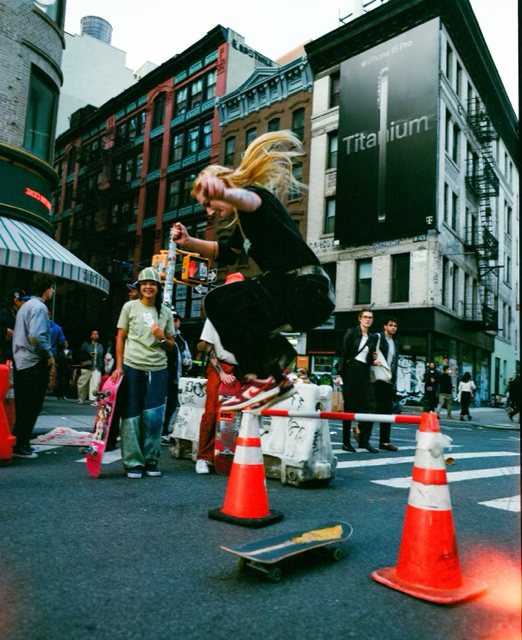A young woman with long blonde hair jumps over orange traffic cones onto a skateboard on a busy city street, surrounded by pedestrians and tall buildings.