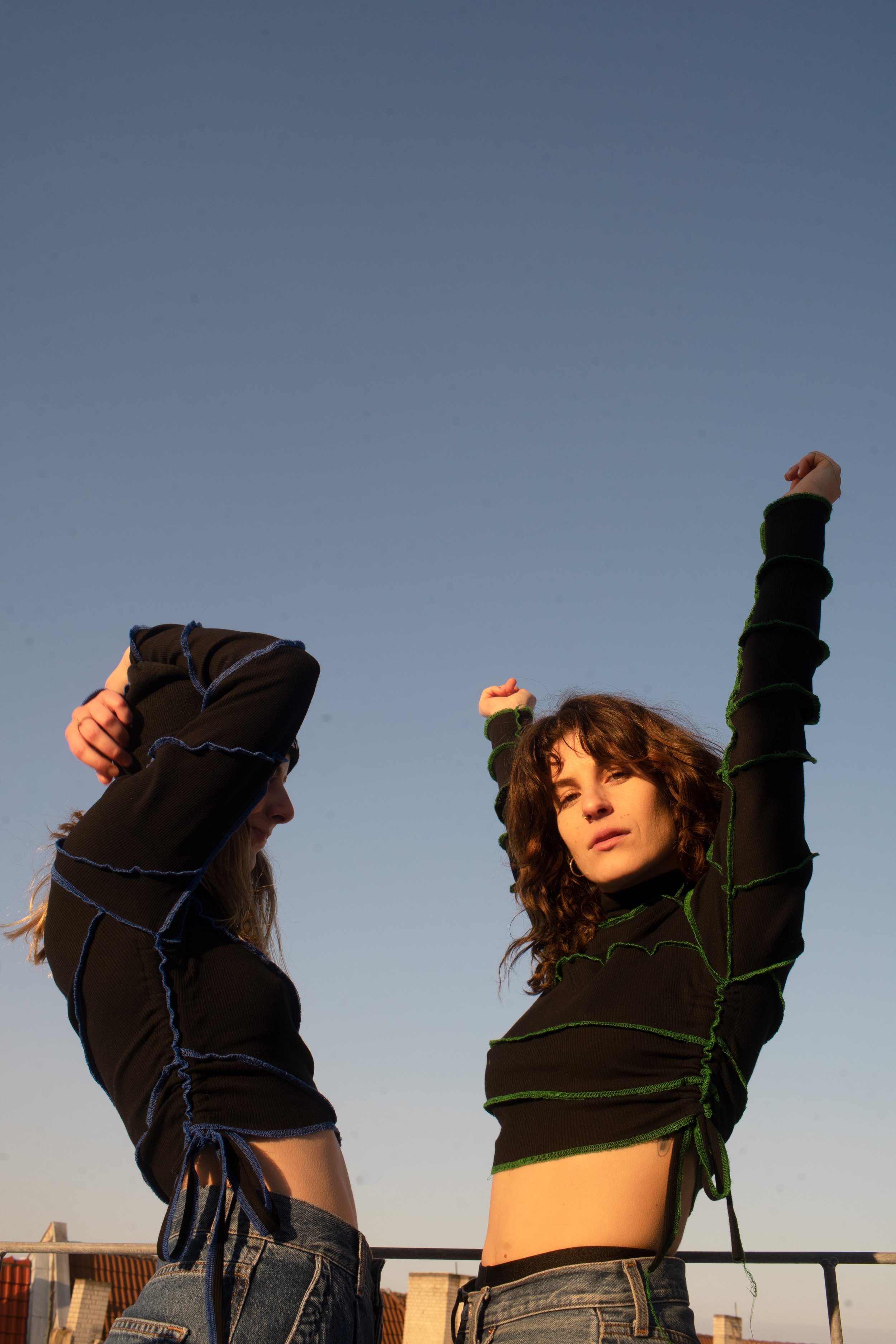 Two women with dark, healthy curls pose outdoors against a clear blue sky, wearing black tops with green and blue stitching and high-waisted jeans.