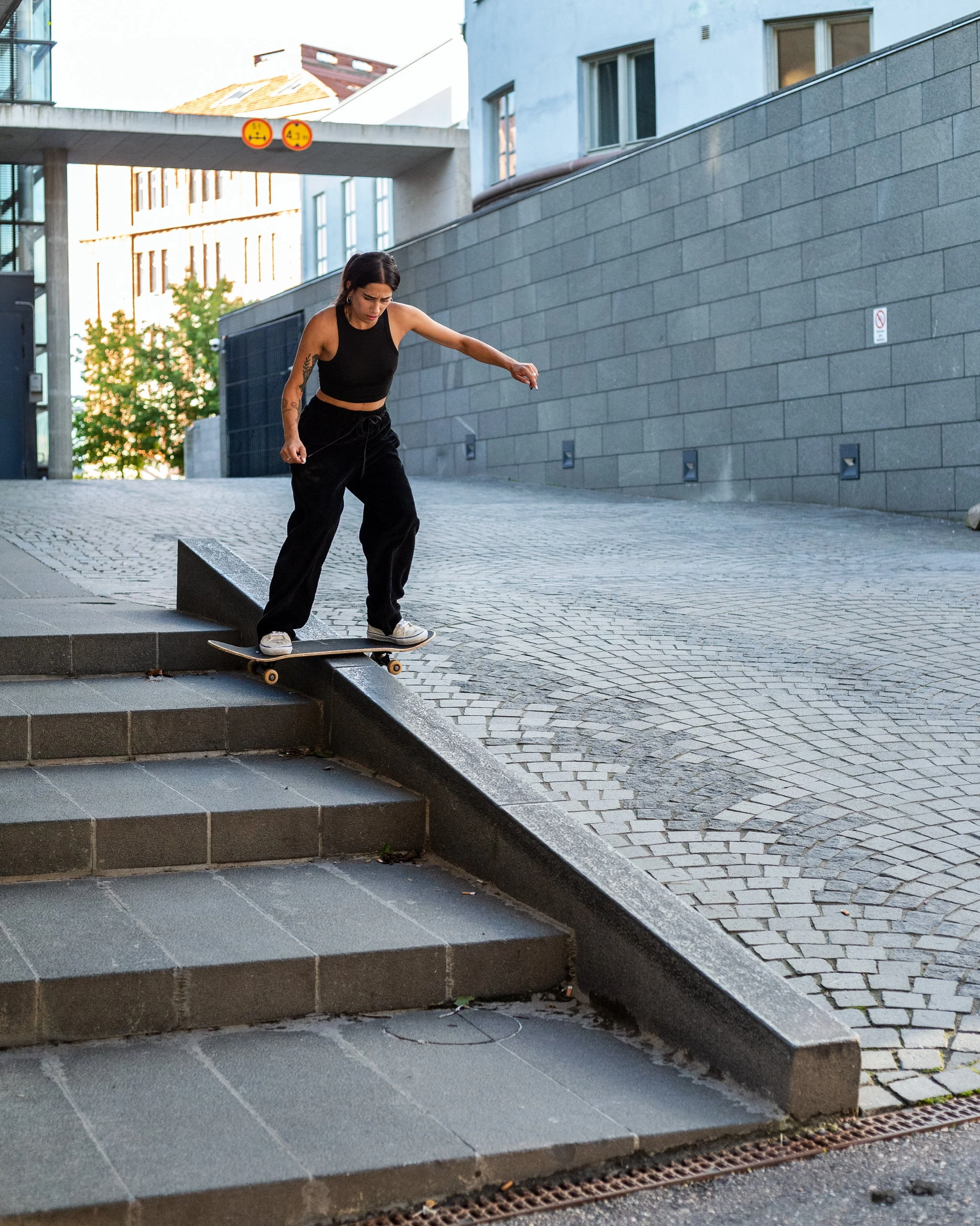 A woman wearing a black tank top and pants skateboarding down a slope with stairs on an urban sidewalk.
