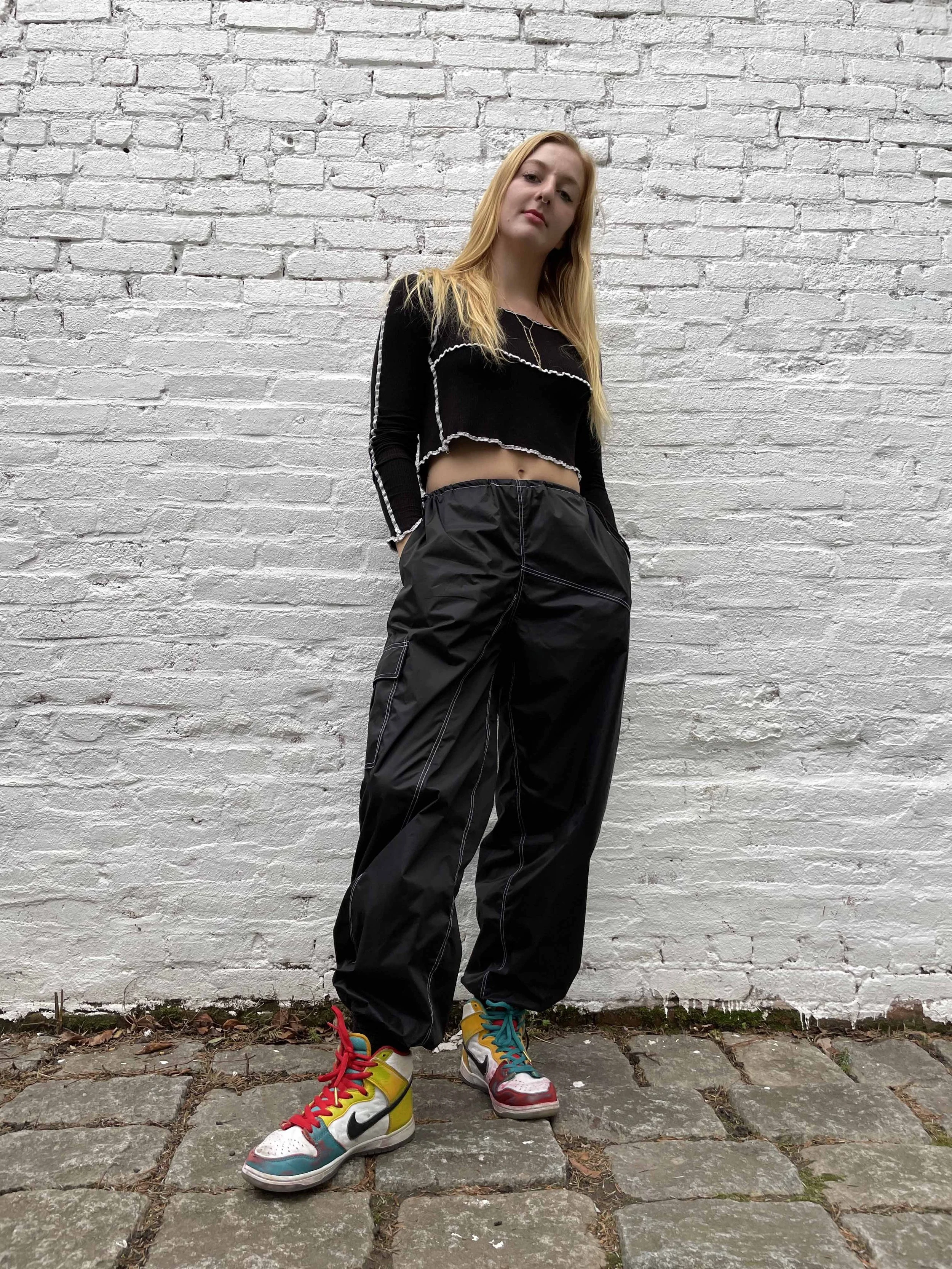 A young woman with long blonde hair stands outdoors against a white brick wall, wearing a black cropped sweater with white trim, black baggy cargo pants, and colorful sneakers.