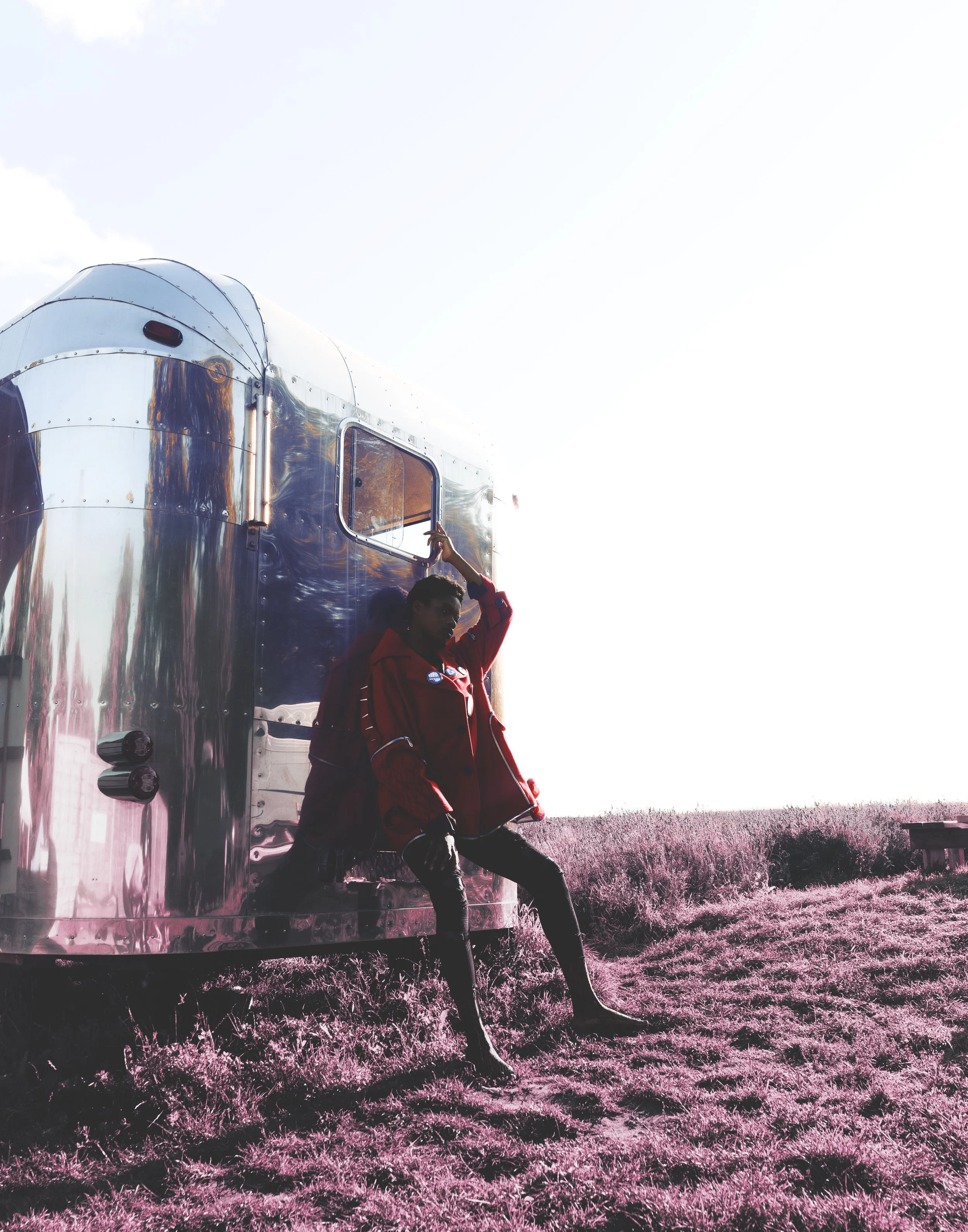Person in a red jacket leaning against a shiny silver trailer in an open field with pink grass, bright sky in the background.
