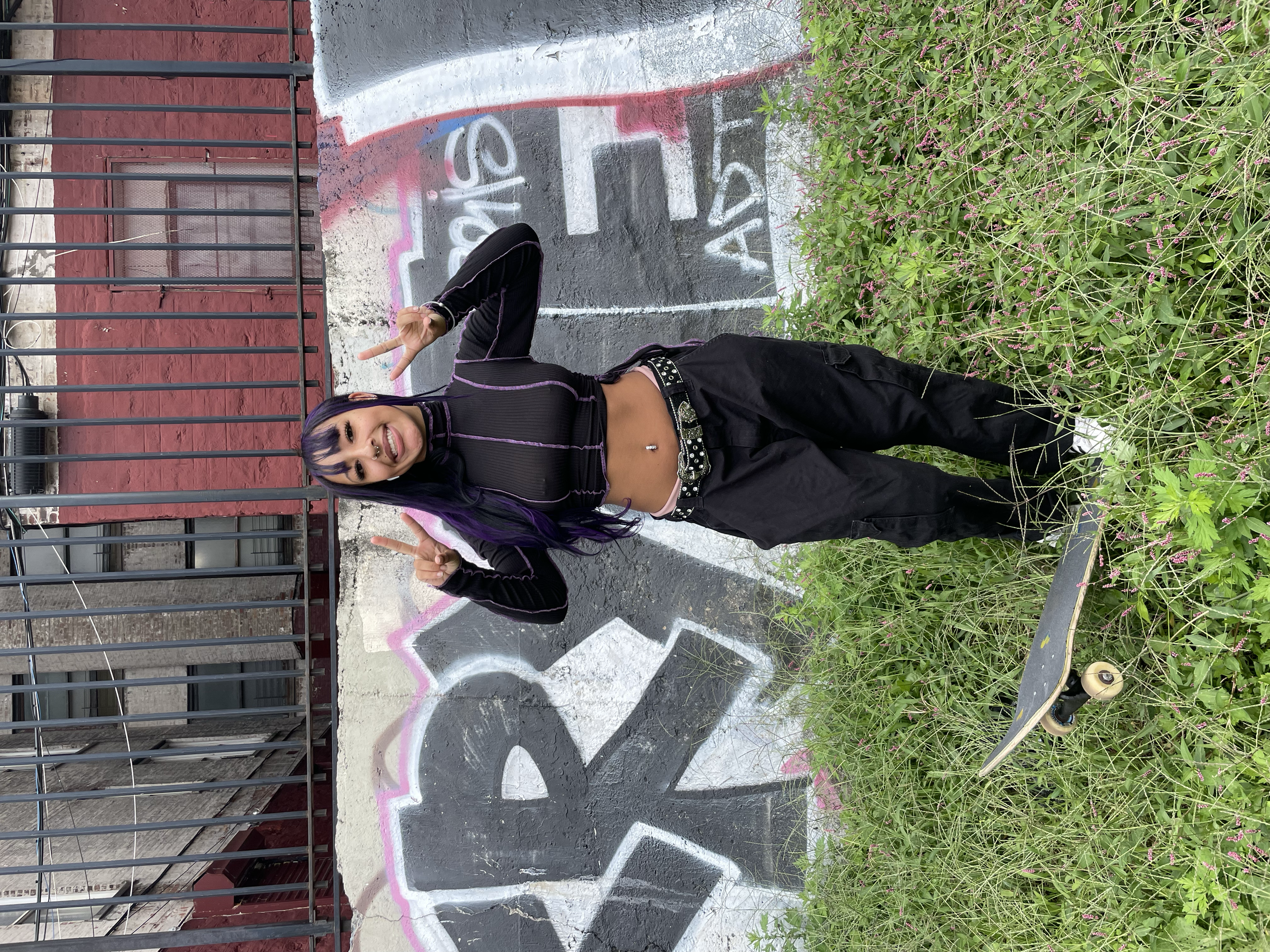 A young woman with purple and black hair posing outdoors in front of a graffiti-covered wall and overgrown plants, holding up peace signs with both hands and smiling.