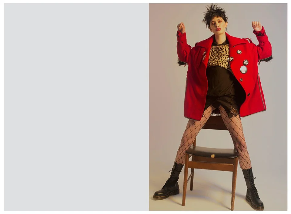 A woman with short, spiky hair standing with legs apart on a chair and arms raised, wearing a red jacket with patches, a leopard print top, fishnet stockings, black shorts, and combat boots, against a plain background.