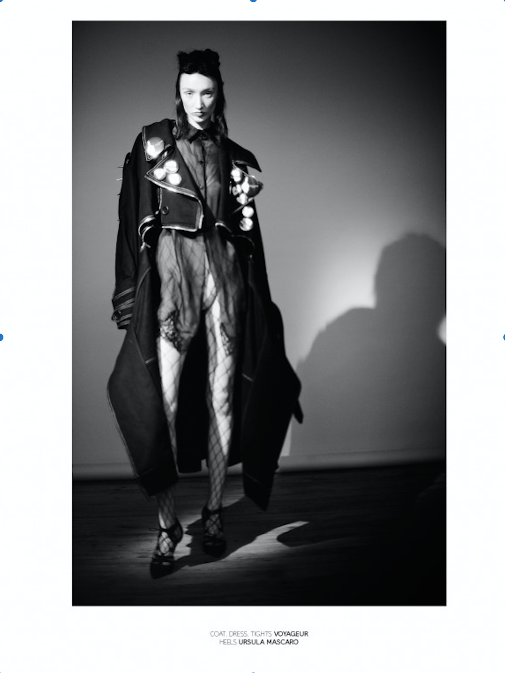 Black and white fashion photo of a woman in a leather jacket decorated with buttons, fishnet tights, high heels, standing against a plain wall.