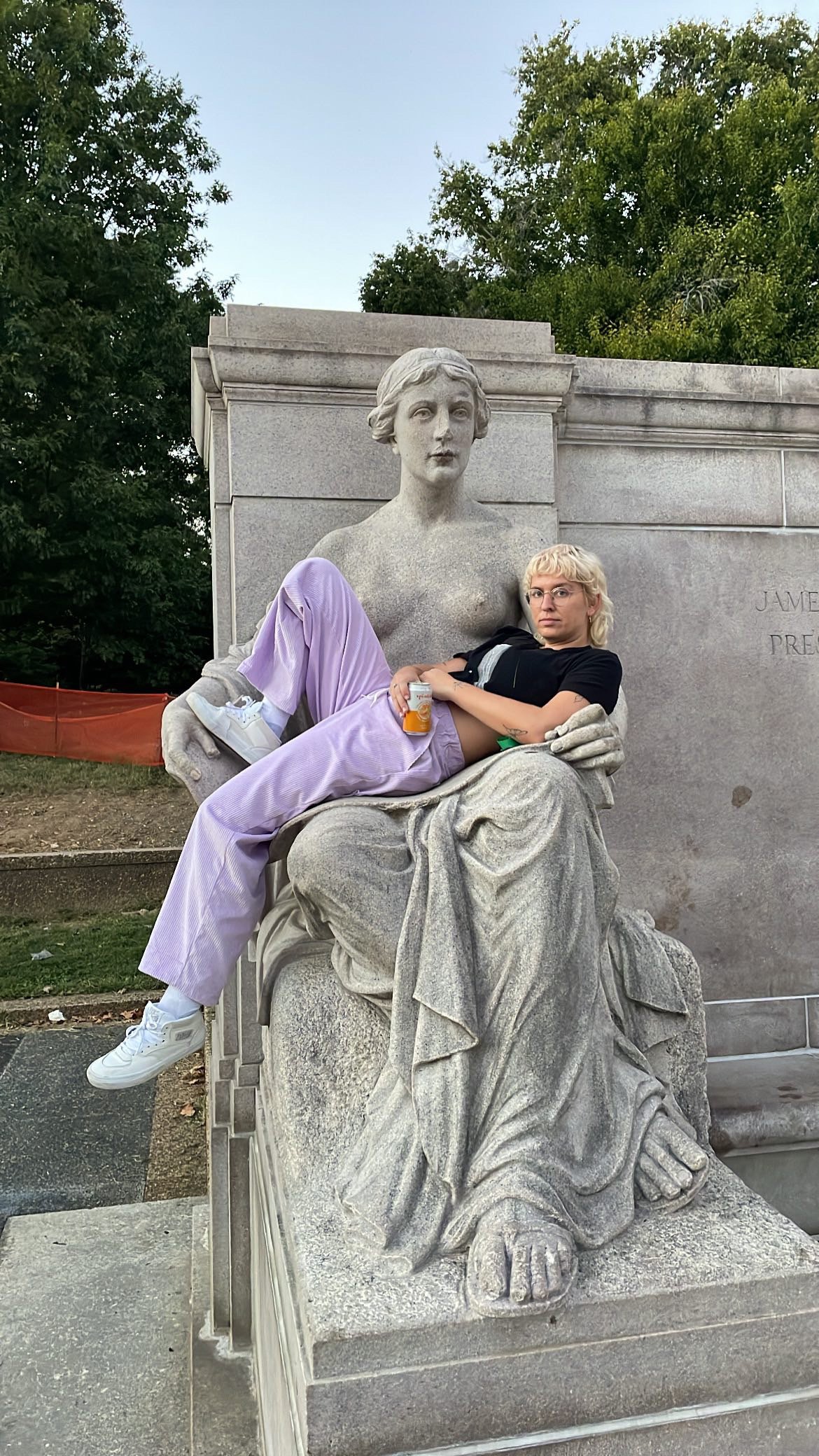 A young woman with blonde hair and glasses sitting on a marble statue of a seated woman in a park, holding a drink and looking at the camera.