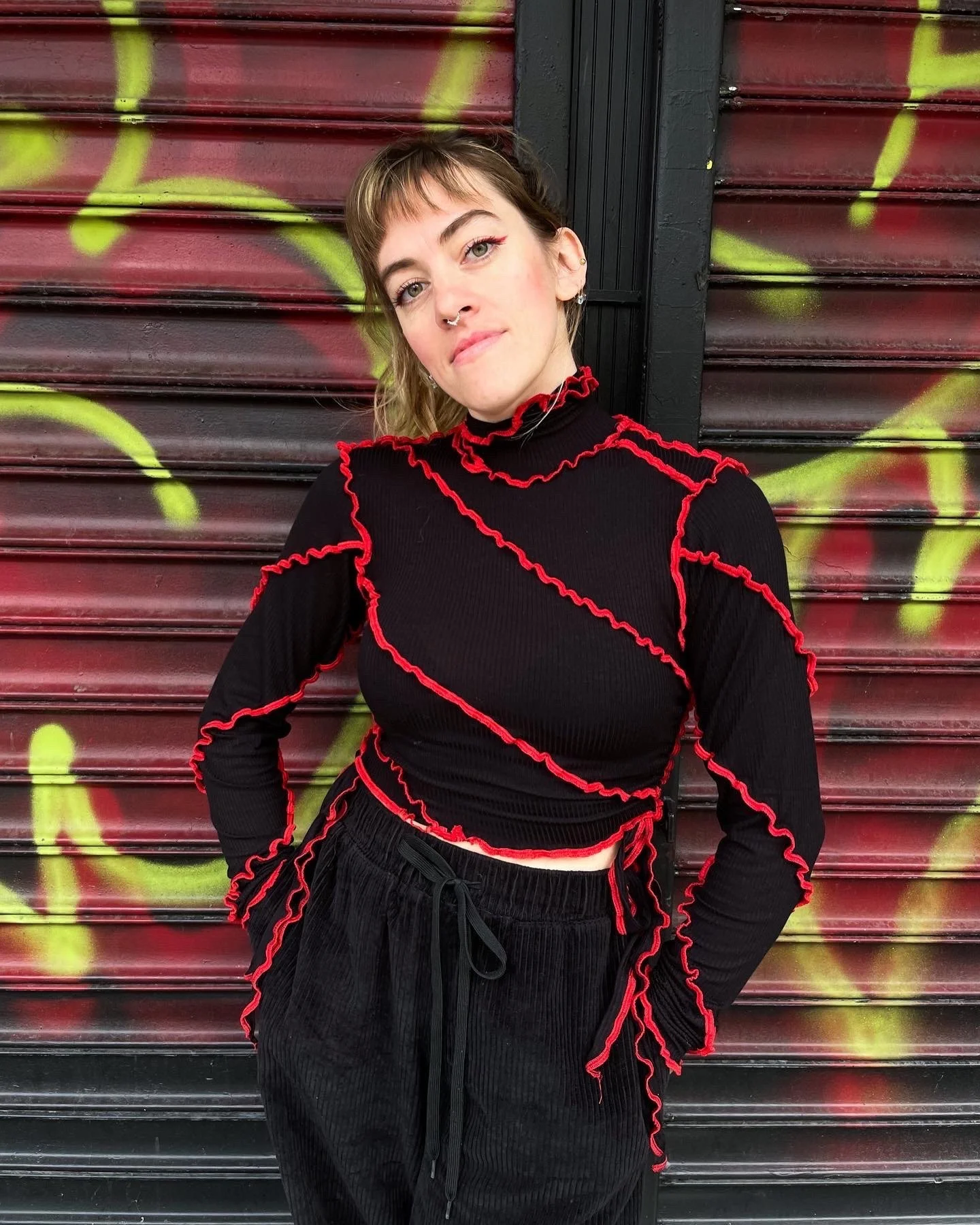 Young woman with short hair and septum piercing standing in front of a graffiti-covered metal shutter, wearing a black ruffled long-sleeve top with red accents and black pants.