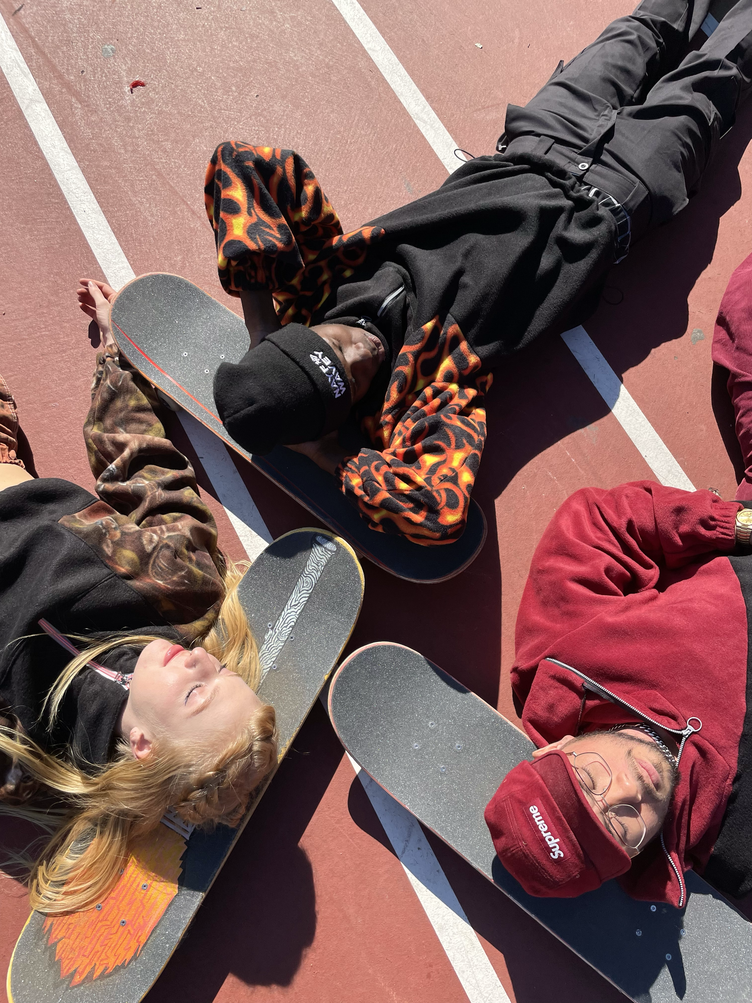 Two young people lying on skateboards on a red outdoor court with white lines, relaxing in the sunlight.