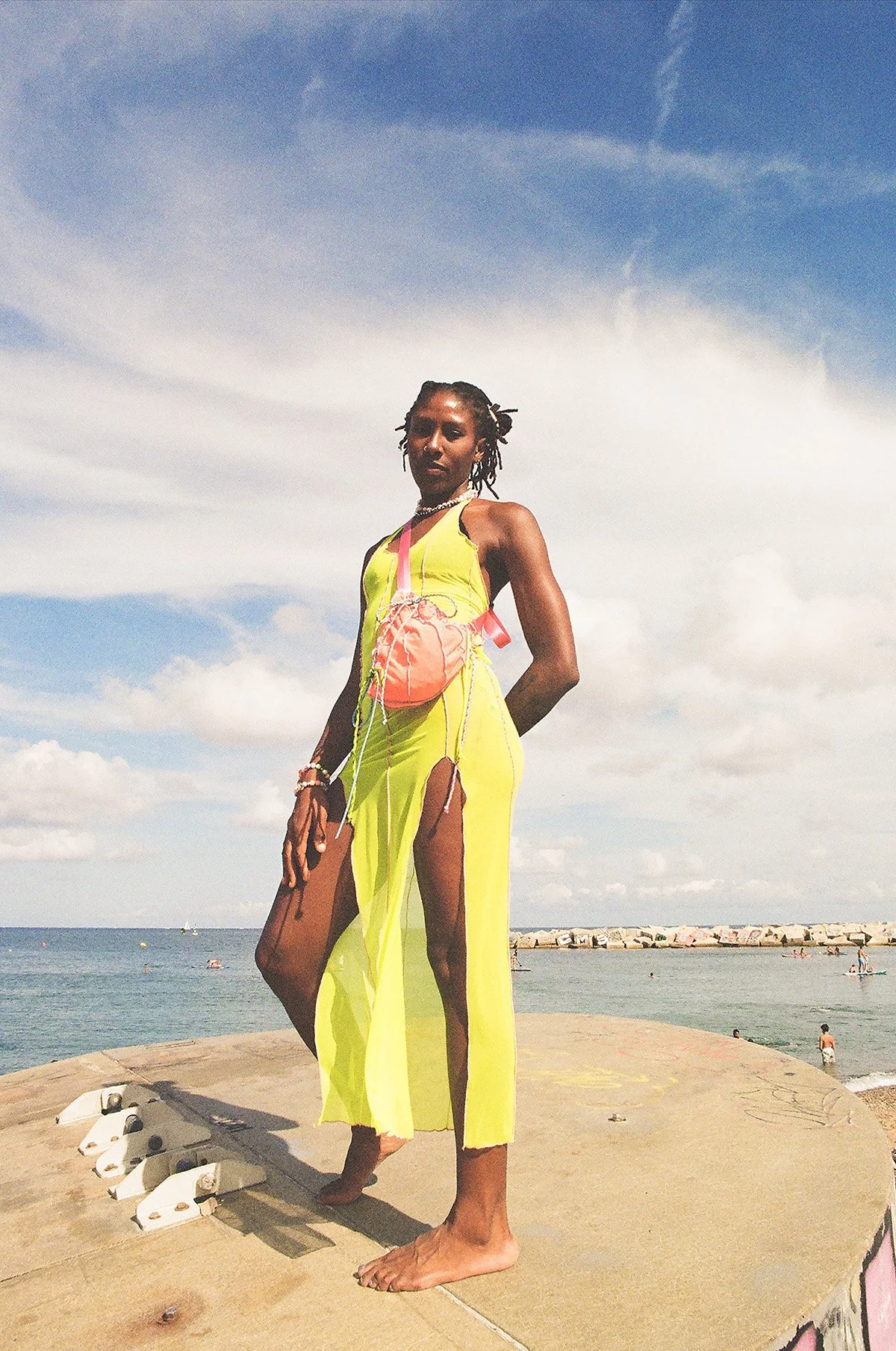 A woman in a yellow maxi dress with high slits stands barefoot on concrete, near the beach, with the ocean and a cloudy sky in the background. She wears a pink purse, layered necklaces, and bracelets.