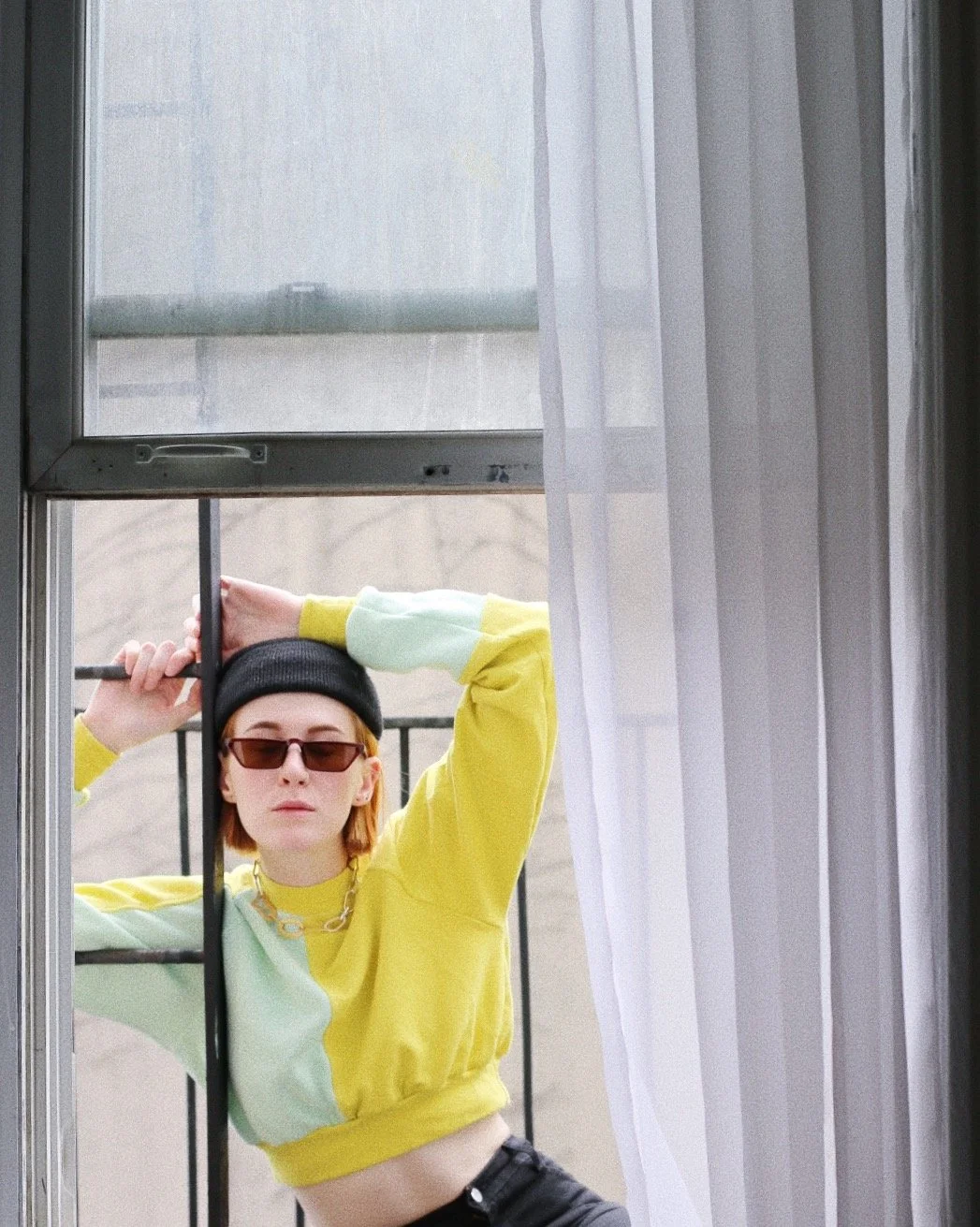 Woman wearing sunglasses, a black beanie, a yellow and mint green color-block sweatshirt, and black pants, standing outside on a balcony, holding onto a window frame, with sheer white curtains and a cloudy sky in the background.