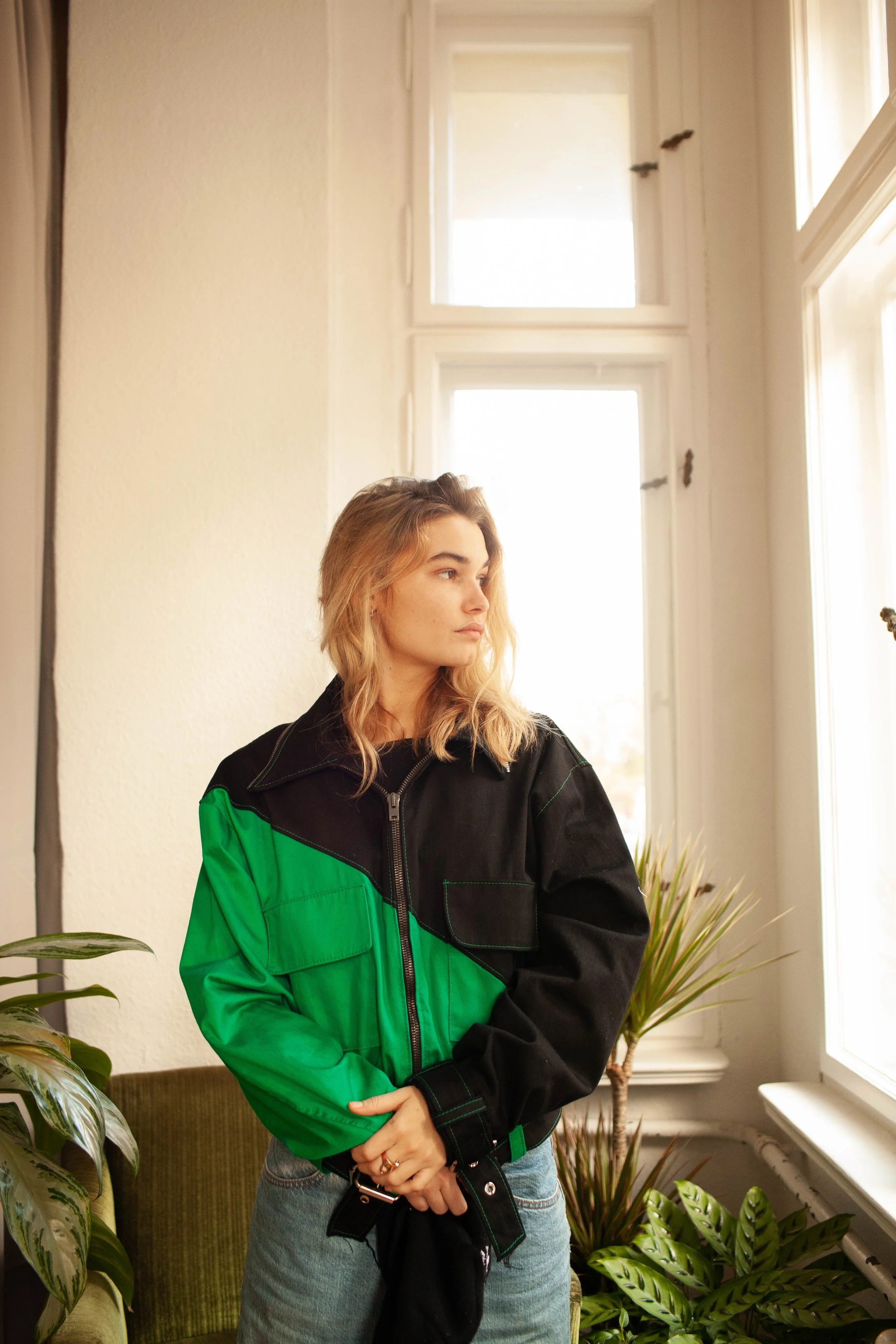 A young woman with blonde hair is standing indoors near a window, wearing a black and green color-blocked jacket and light blue jeans, with plants and a green chair in the background.