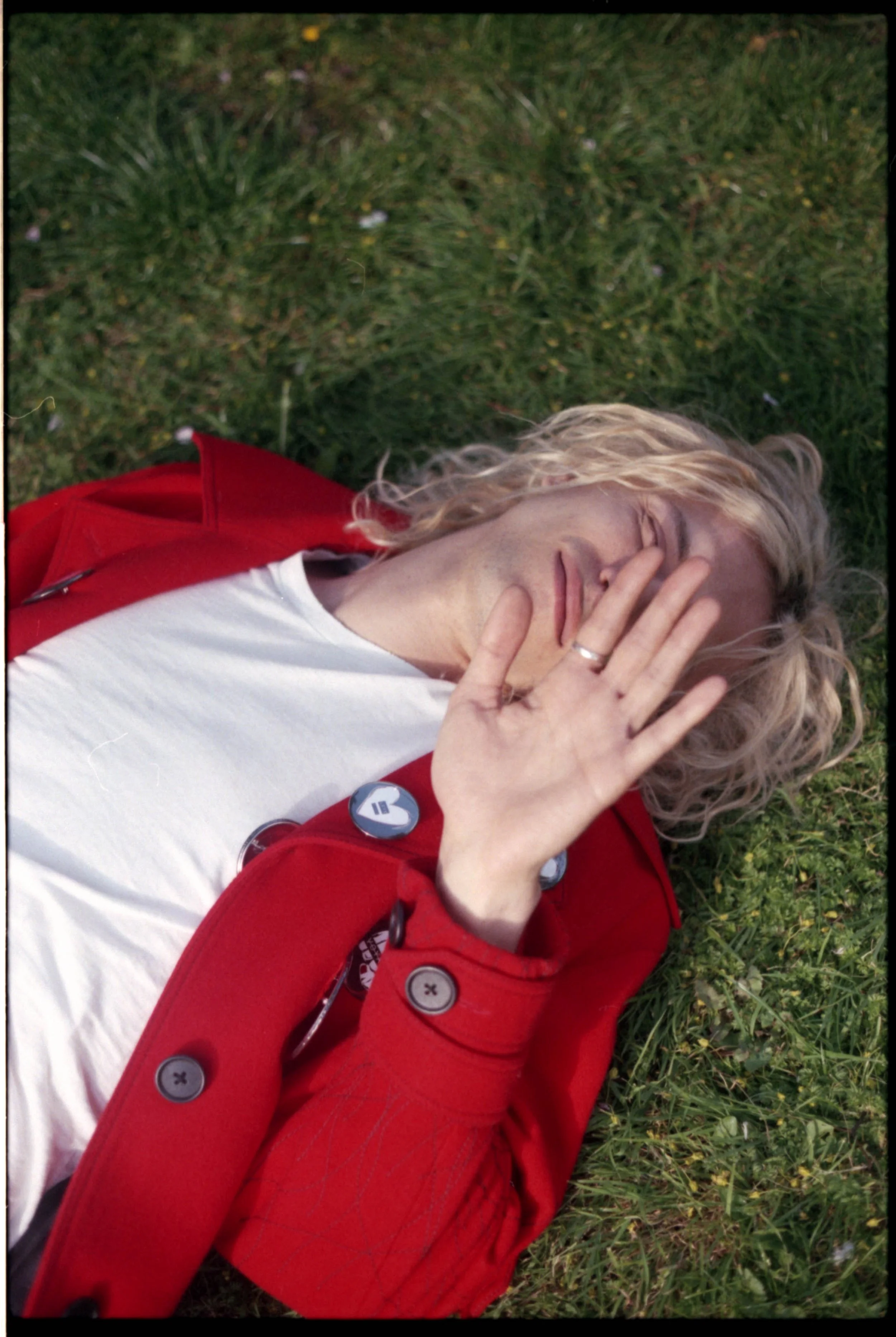 A person with long, curly blonde hair lying on grass outdoors, wearing a red jacket and a white shirt, covering part of their face with their hand.