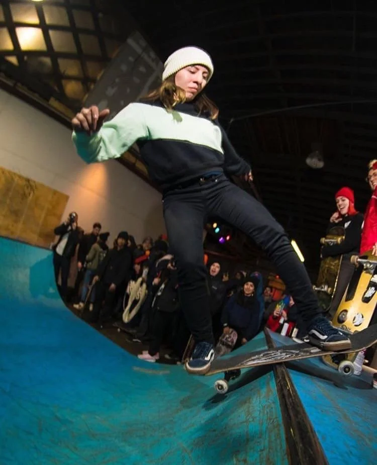 Young woman in a white beanie and green and black sweatshirt doing a skateboarding trick at an indoor skatepark, surrounded by spectators.