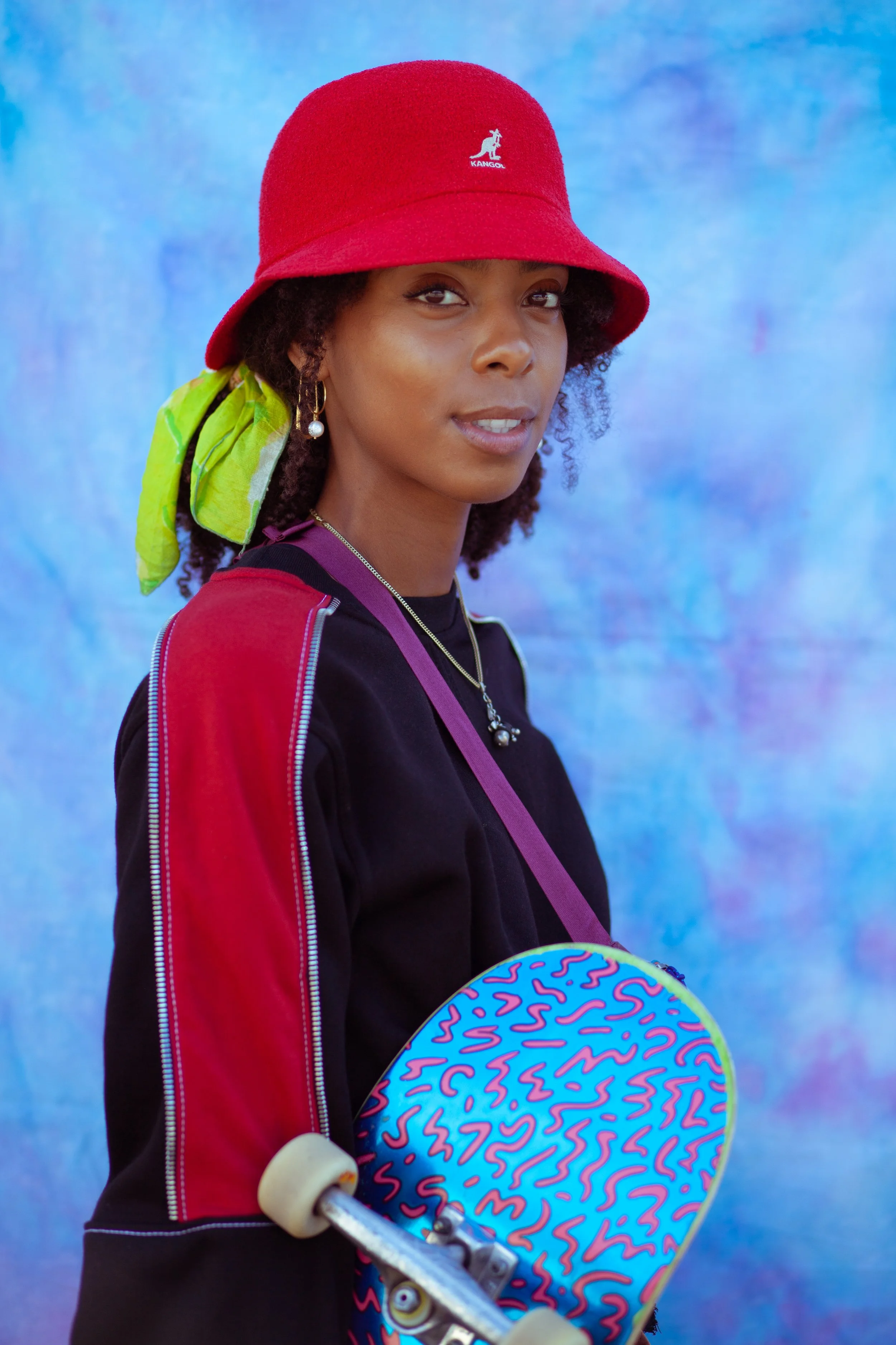 A young woman wearing a red Kangol bucket hat, black and red jacket, and a green scarf tied around her hair. She has earrings, a necklace, and a purple crossbody bag. She is holding a skateboard with a colorful, abstract pattern on its deck and white