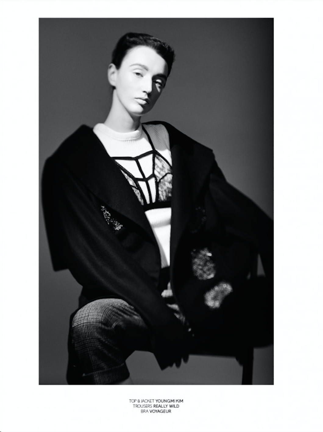 Black and white fashion photograph of a woman wearing a top and jacket, patterned trousers, and a decorative bra visible under her open jacket, sitting against a plain background.