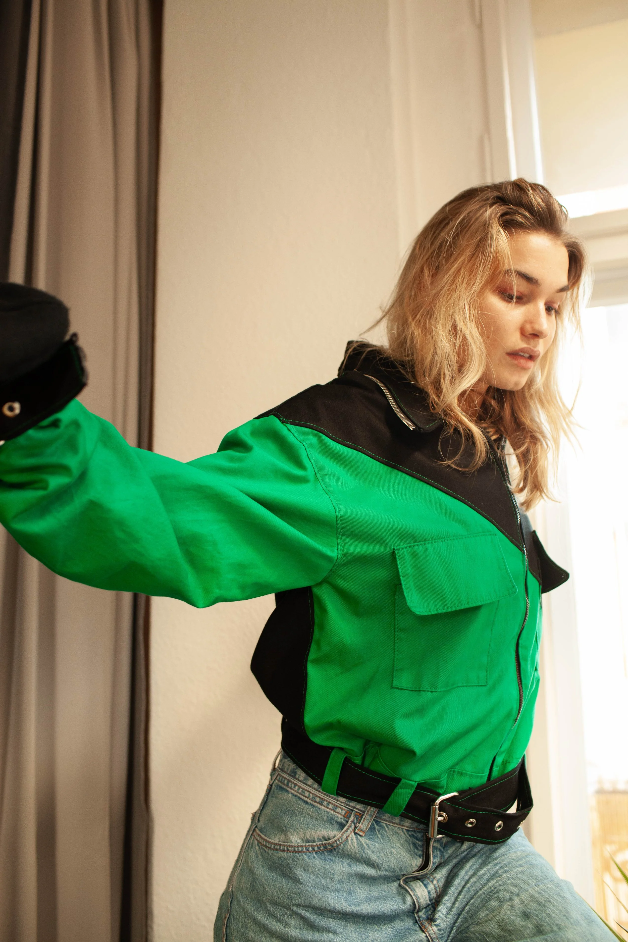 A young woman with blonde wavy hair wearing a green and black jacket and light blue jeans, standing indoors near a window.