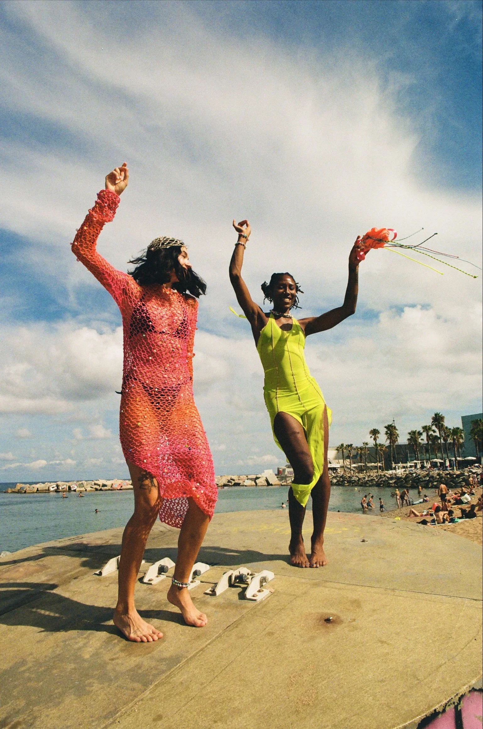 Two women dancing on a concrete platform by the beach, with a body of water, people in the background, and a sky with clouds.