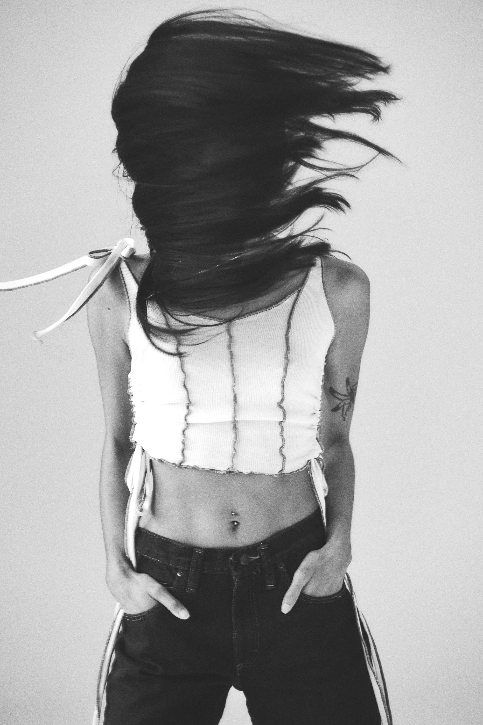 A woman with long dark hair flipping her hair, wearing a white sleeveless top with vertical stripes and dark jeans, standing against a plain background.