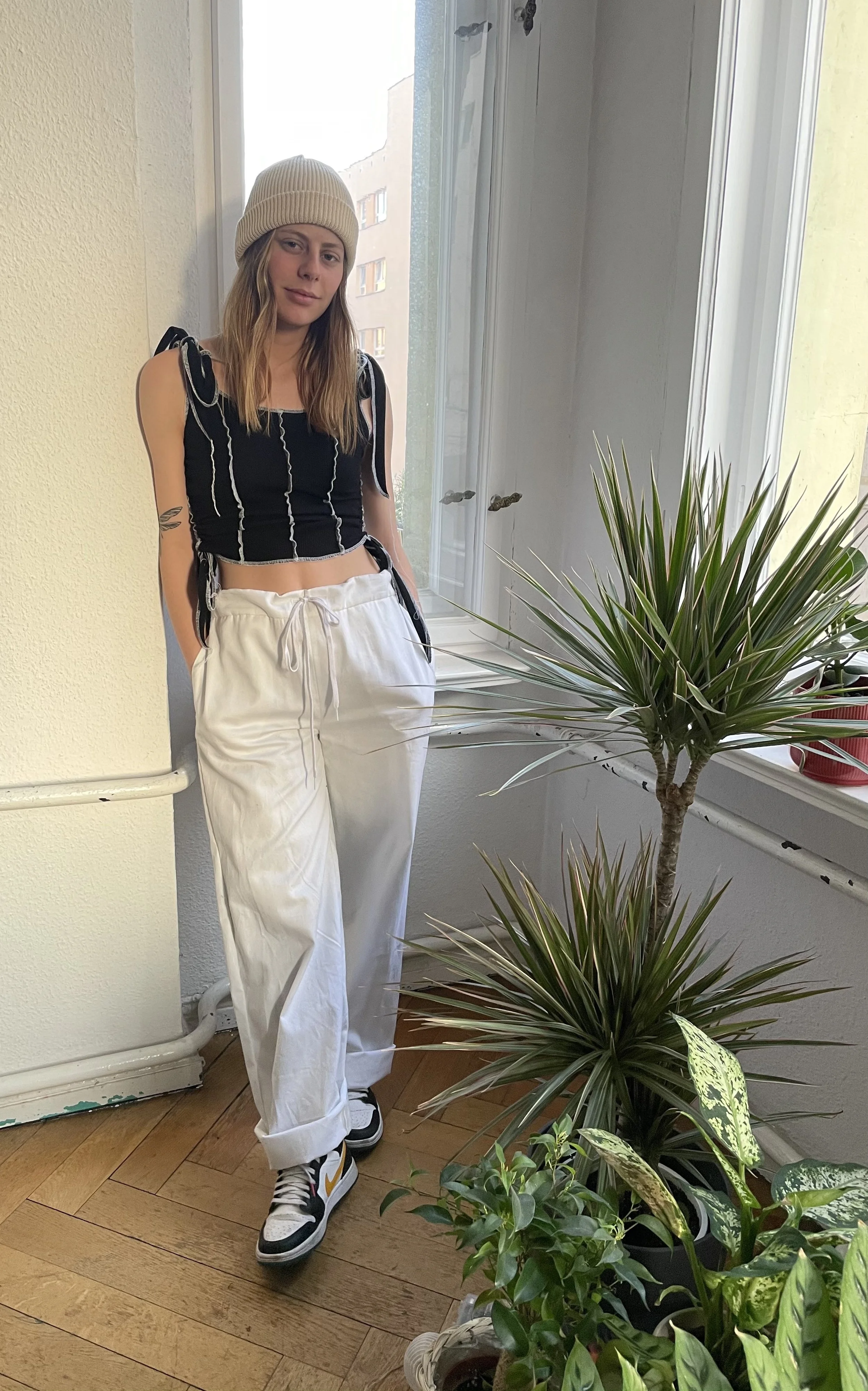 A young woman in casual black and white outfit standing inside near large window and potted plants.