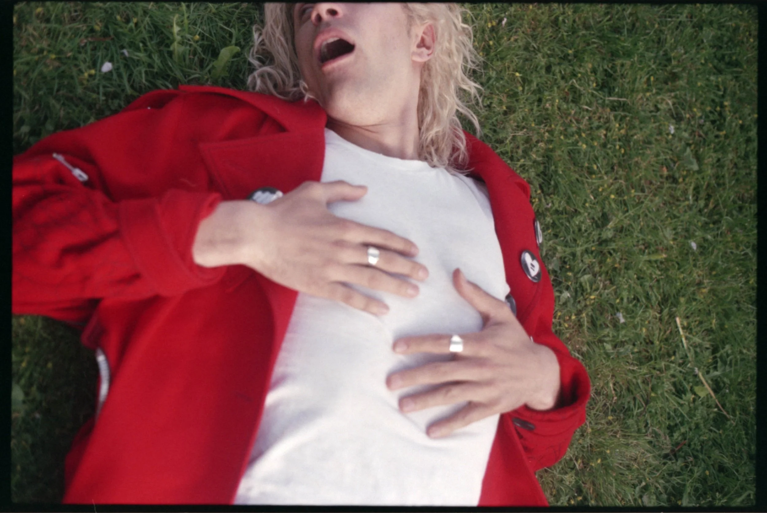 A person lying on grass wearing a red jacket and a white shirt, holding their chest with both hands, with long blond curly hair and an expressive facial expression.