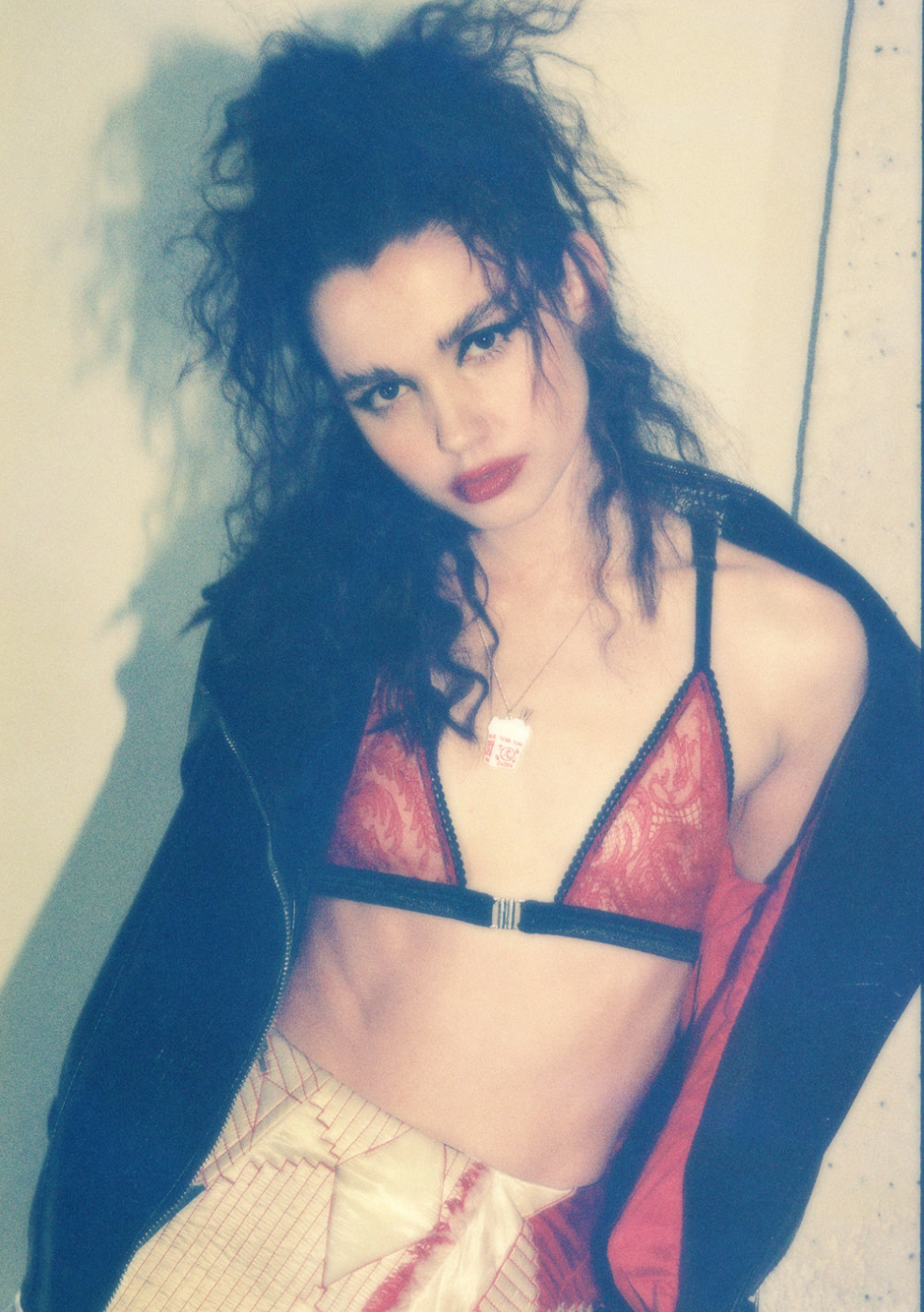 Woman with curly dark hair wearing red lace lingerie top, black jacket off the shoulders, and a necklace with a dog tag pendant, posing against a plain background.