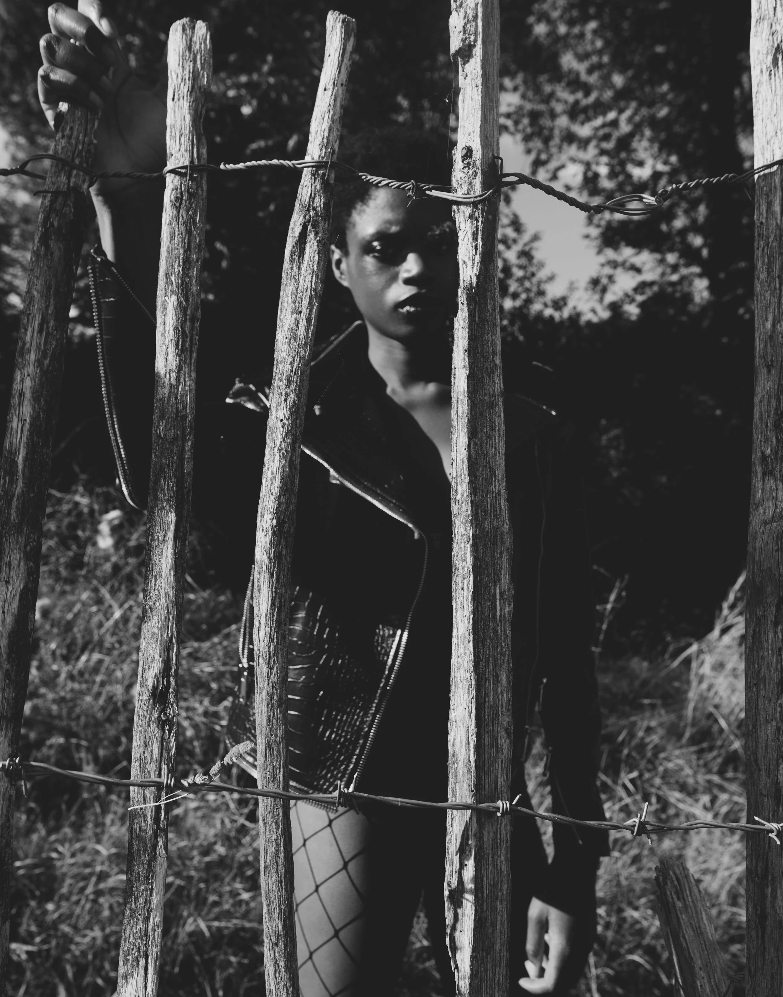 A woman with short hair, wearing a leather jacket and fishnet tights, stands behind a weathered wooden fence with barbed wire, outdoors against a background of trees and grass, captured in black and white.