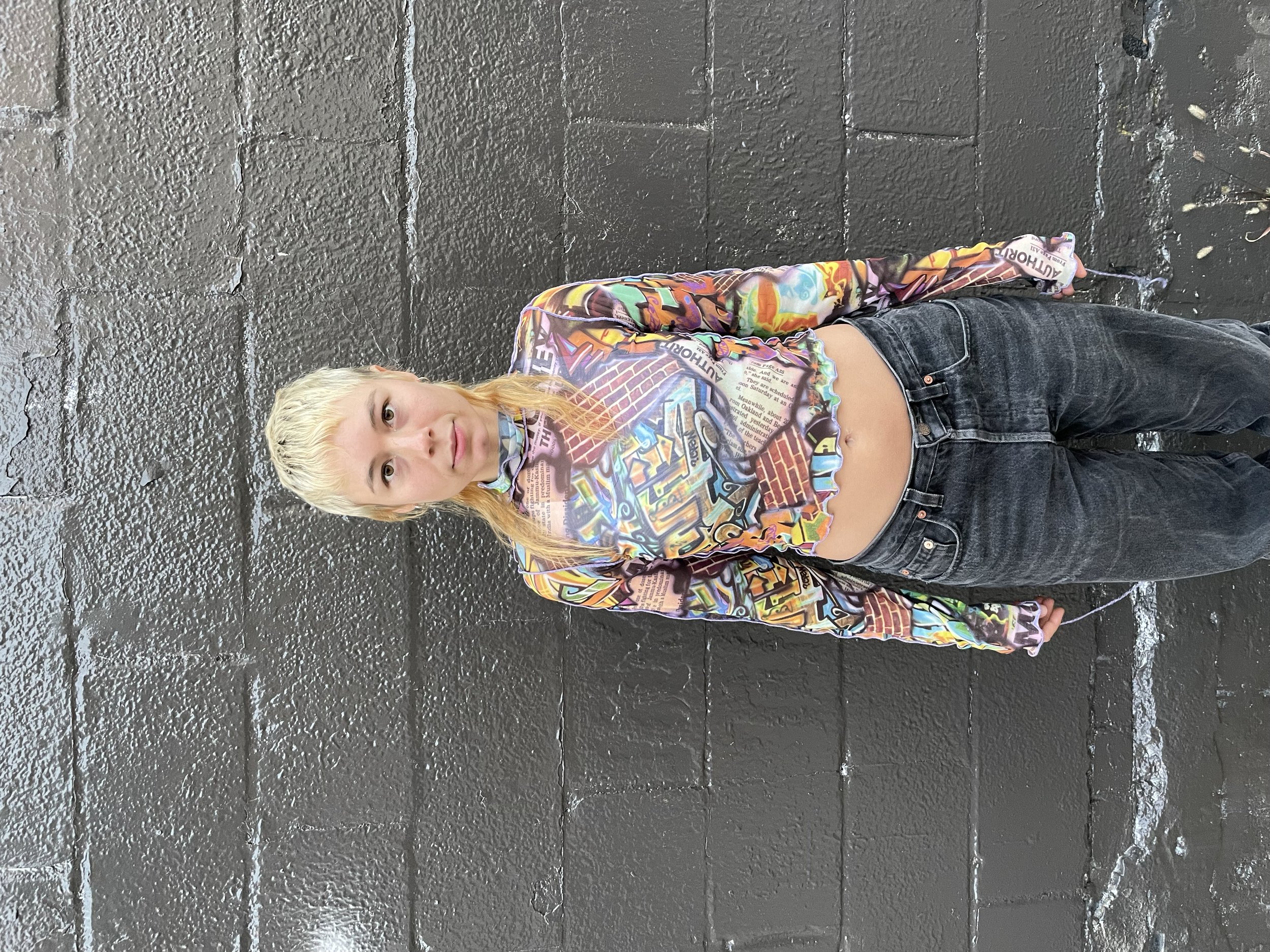 A young girl with blonde and dyed black hair, wearing a colorful graffiti-style long-sleeve shirt and black jeans, standing against a black brick wall.