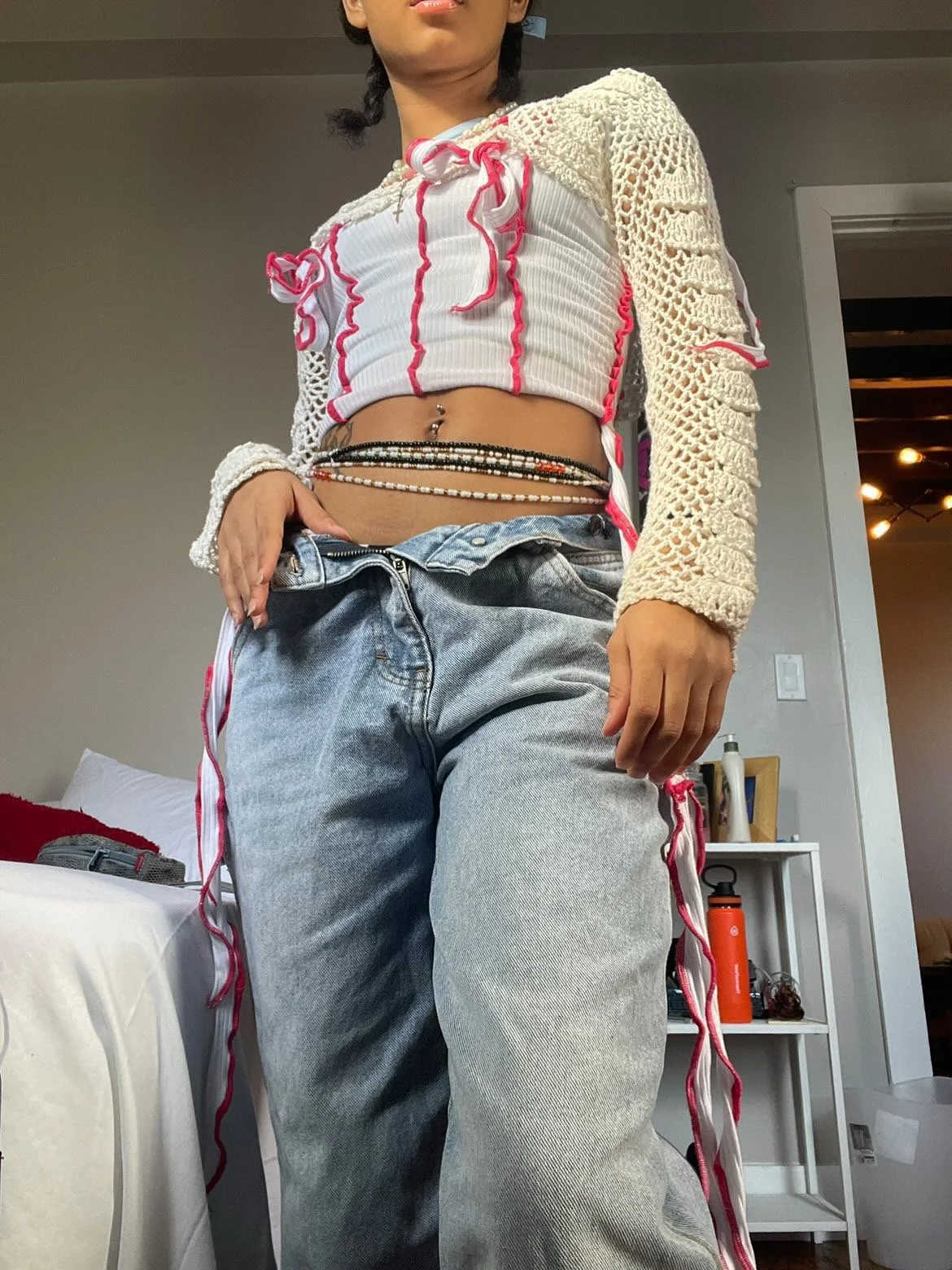 A woman is standing indoors wearing a white cropped top with red frills, loose high-waisted jeans, and a crochet cardigan with long sleeves. She has a belly button piercing and a beaded waist chain. Part of her face and hair are visible; her hair is 