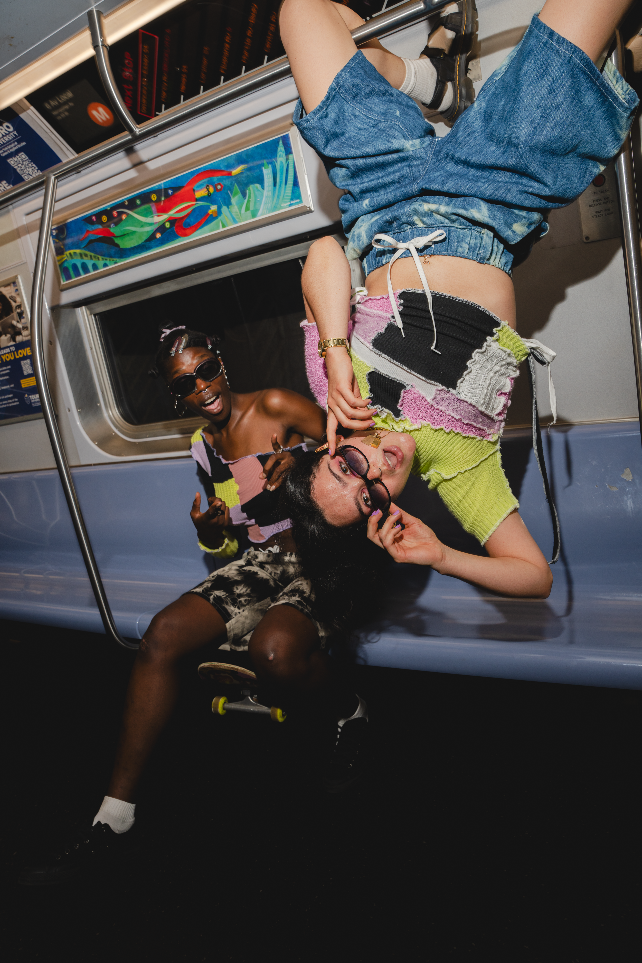 Three young women hanging upside down inside a subway train, smiling and having fun. They are wearing colorful, trendy outfits and sunglasses.