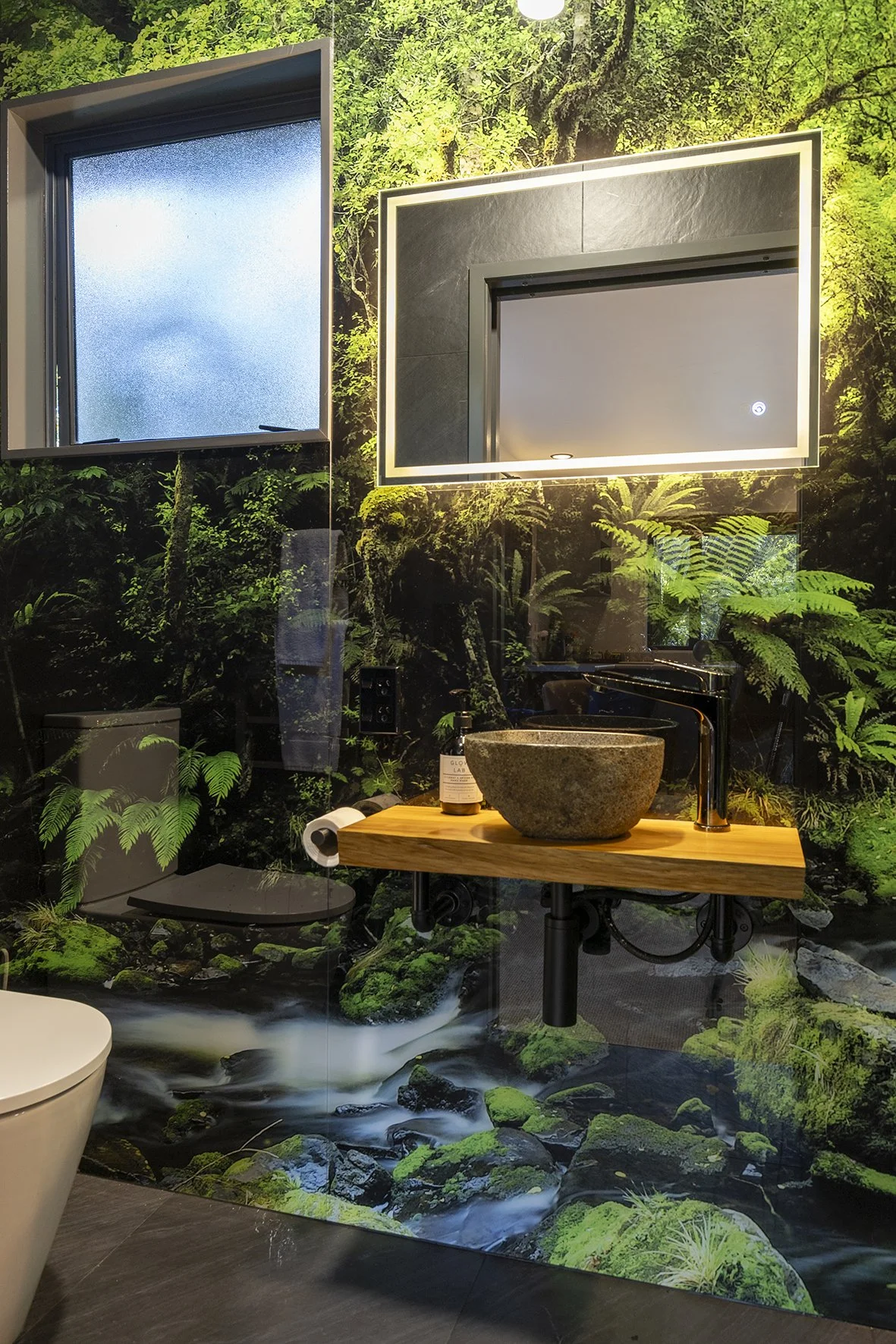 A bathroom with a nature-themed mural of a lush green forest and flowing stream, featuring a wooden countertop with a stone sink and a black faucet. There is a mirror with illuminated border, a frosted window, a toilet, and a bottle of hand soap visi