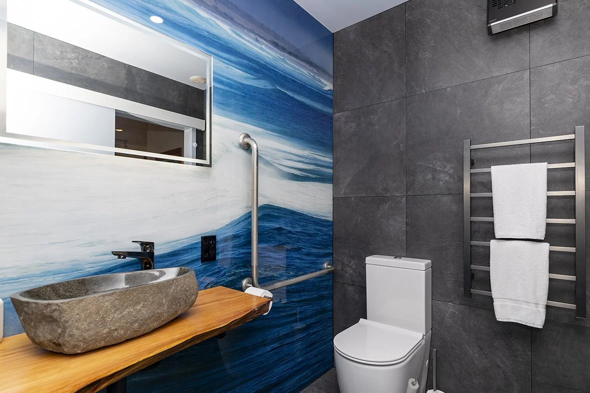 Modern bathroom with a mural of ocean waves on the wall, a stone sink on a wooden countertop, a white toilet, and a metal towel rack with towels.