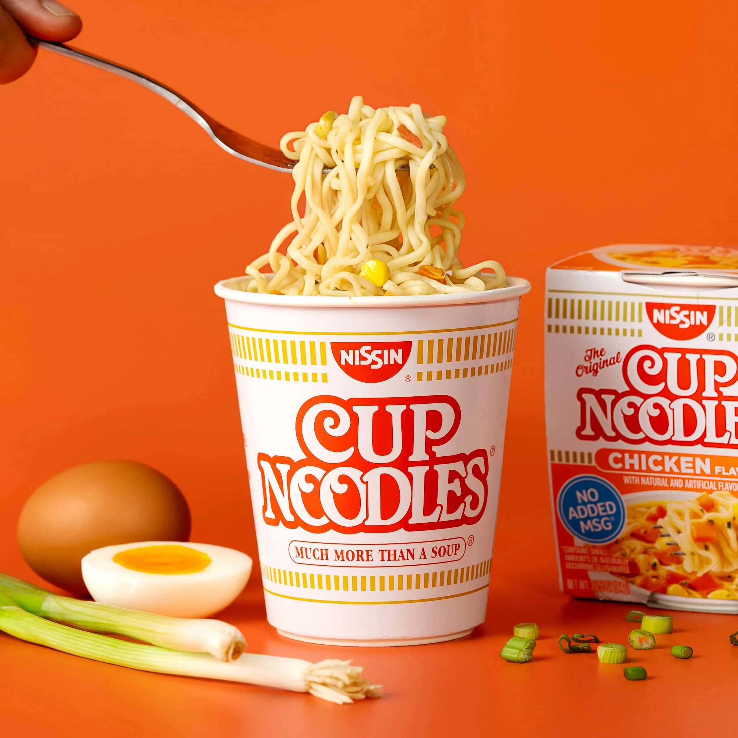A cup of Nissin Cup Noodles with noodles being lifted by a fork, along with a soft-boiled egg, a whole egg, green onions, and a box of Chicken flavor Cup Noodles on an orange background.