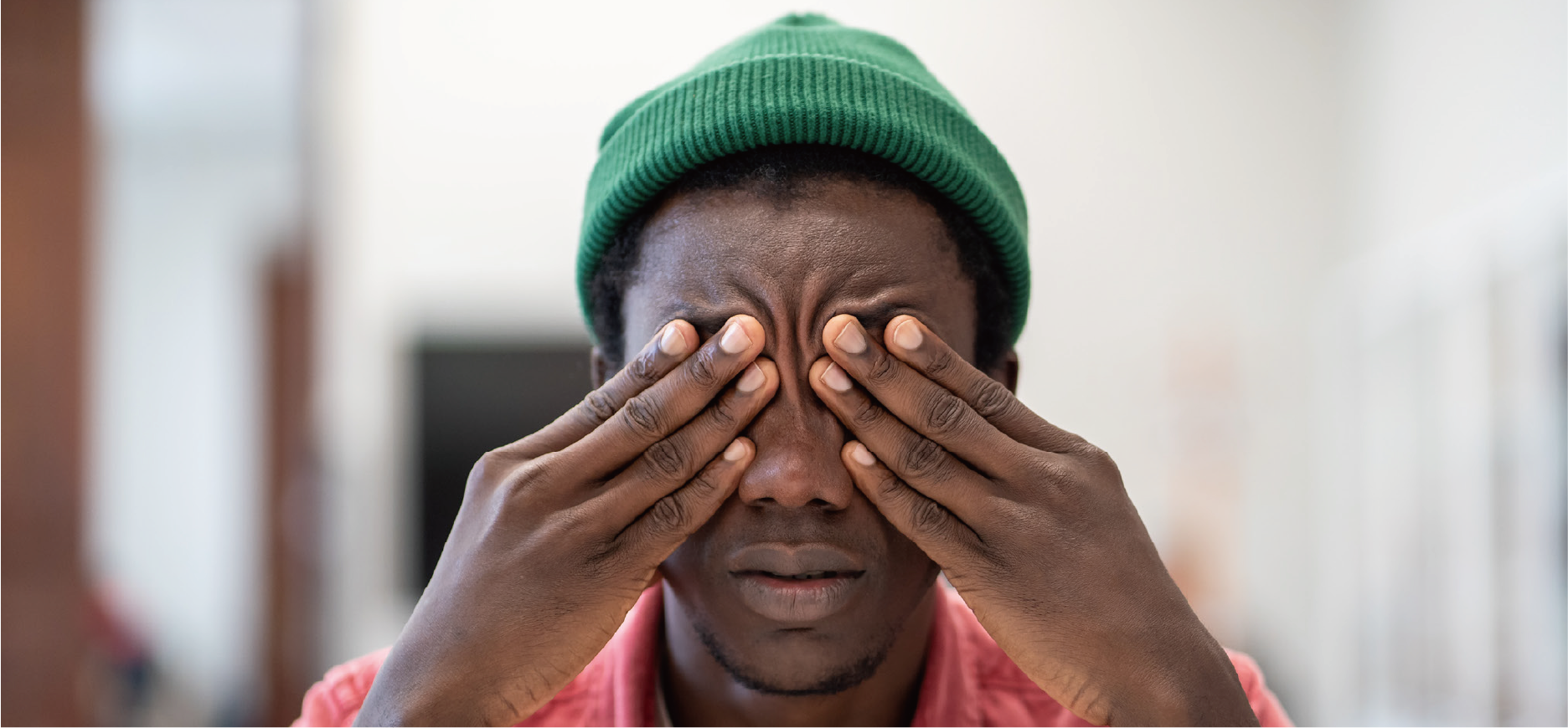 A man wearing a green beanie and red shirt, with his eyes closed and hands covering his eyes, appearing to be in distress or deep thought.