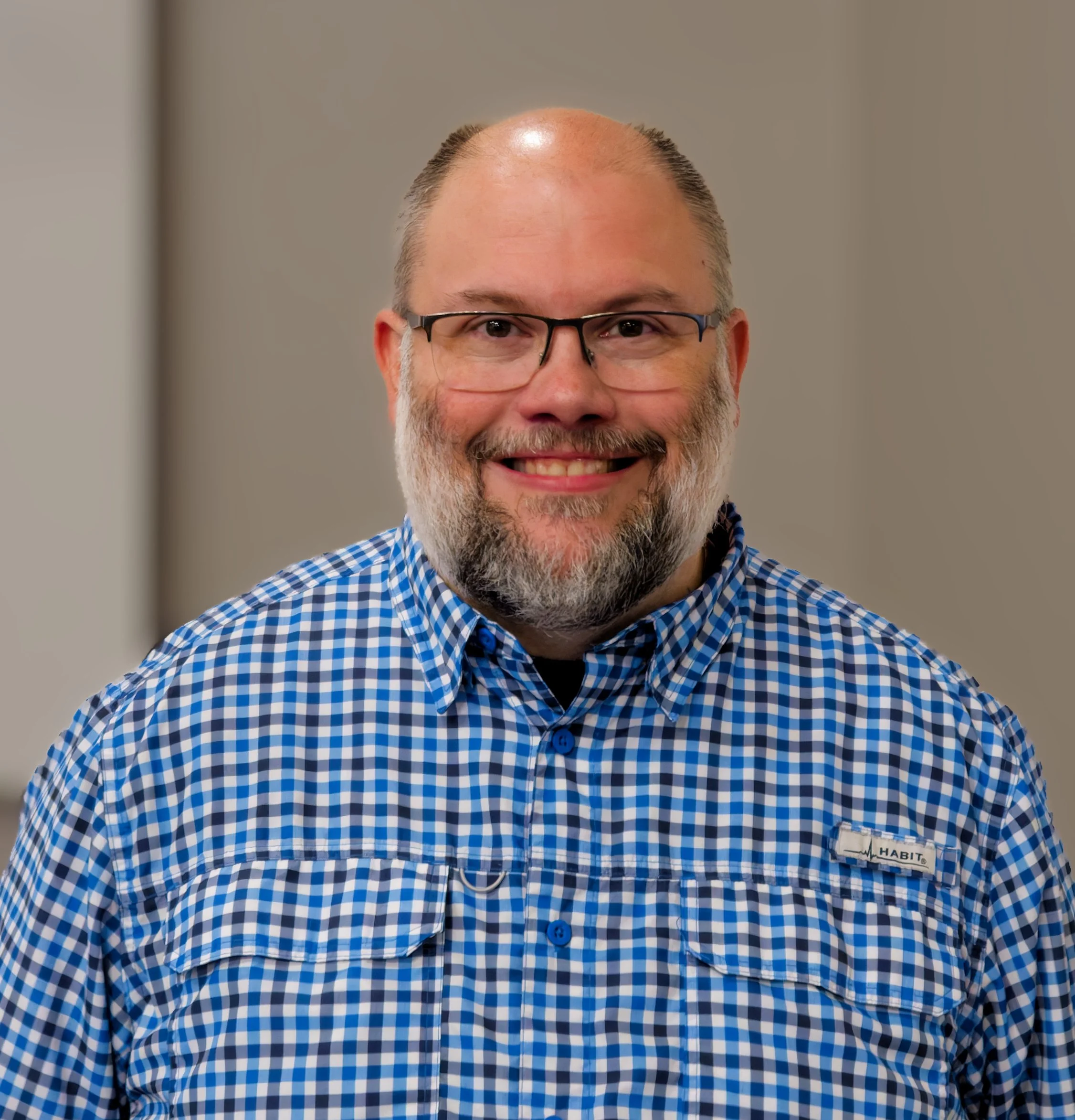A smiling middle-aged man with a beard and bald head, wearing a white button-up shirt, outdoors with a blurred green background.