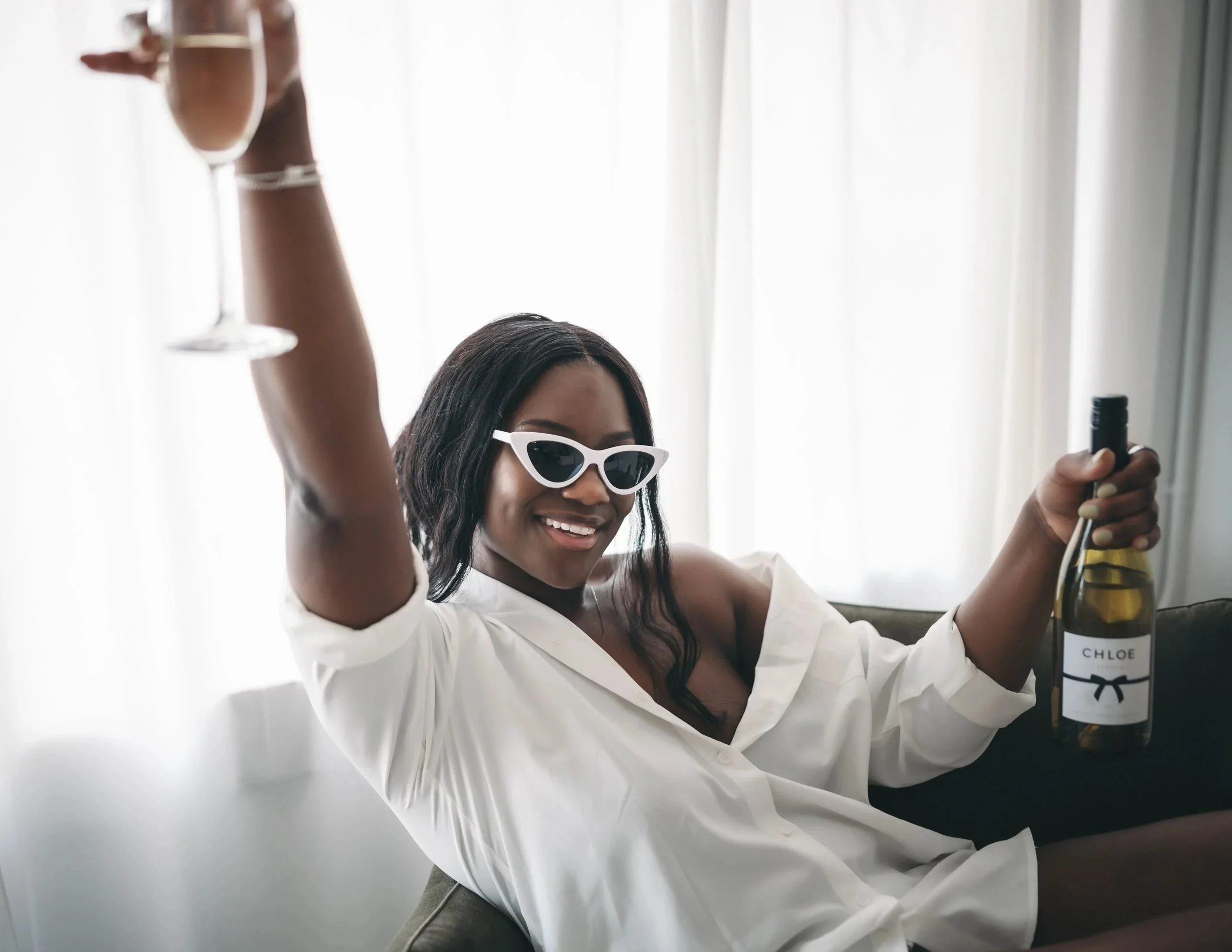 Young woman wearing sunglasses and a white shirt smiling, raising a champagne glass, and holding a bottle of Chloe wine in a bright room with sheer curtains.