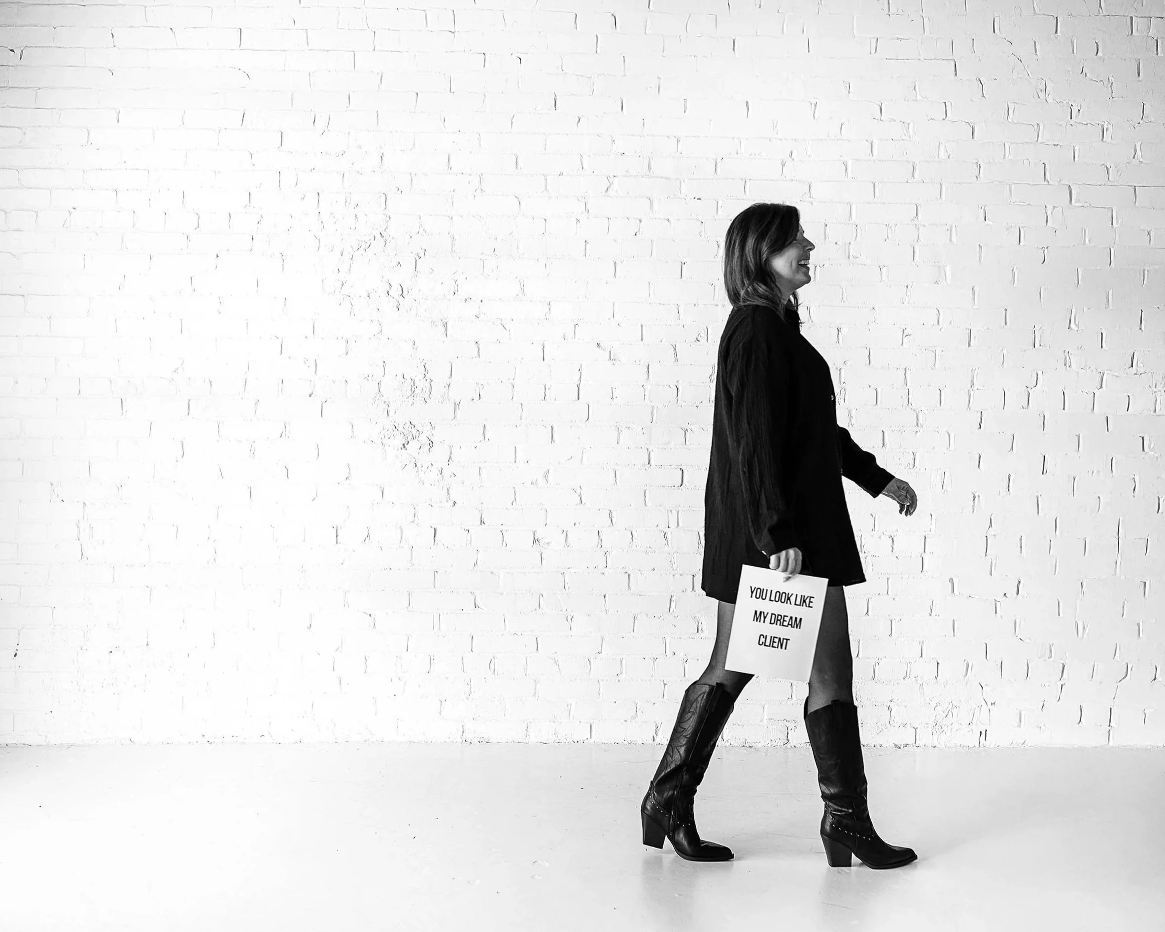 A woman in black leather boots and a black jacket, smiling, holding a sign that reads 'You look like my dream client,' standing in front of a white brick wall.