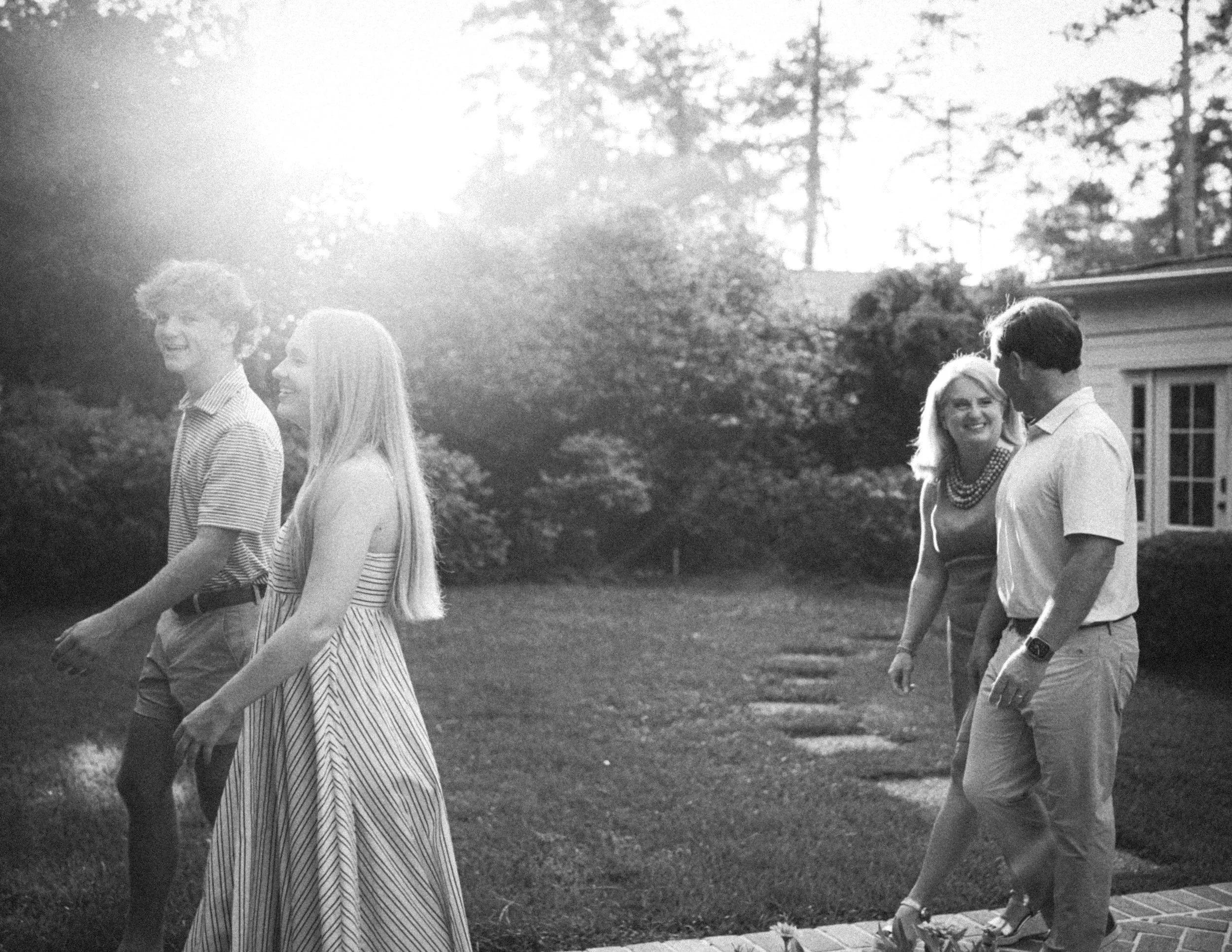 Four young adults walking outdoors on a grassy area, smiling and chatting, with trees and a house in the background during daytime.