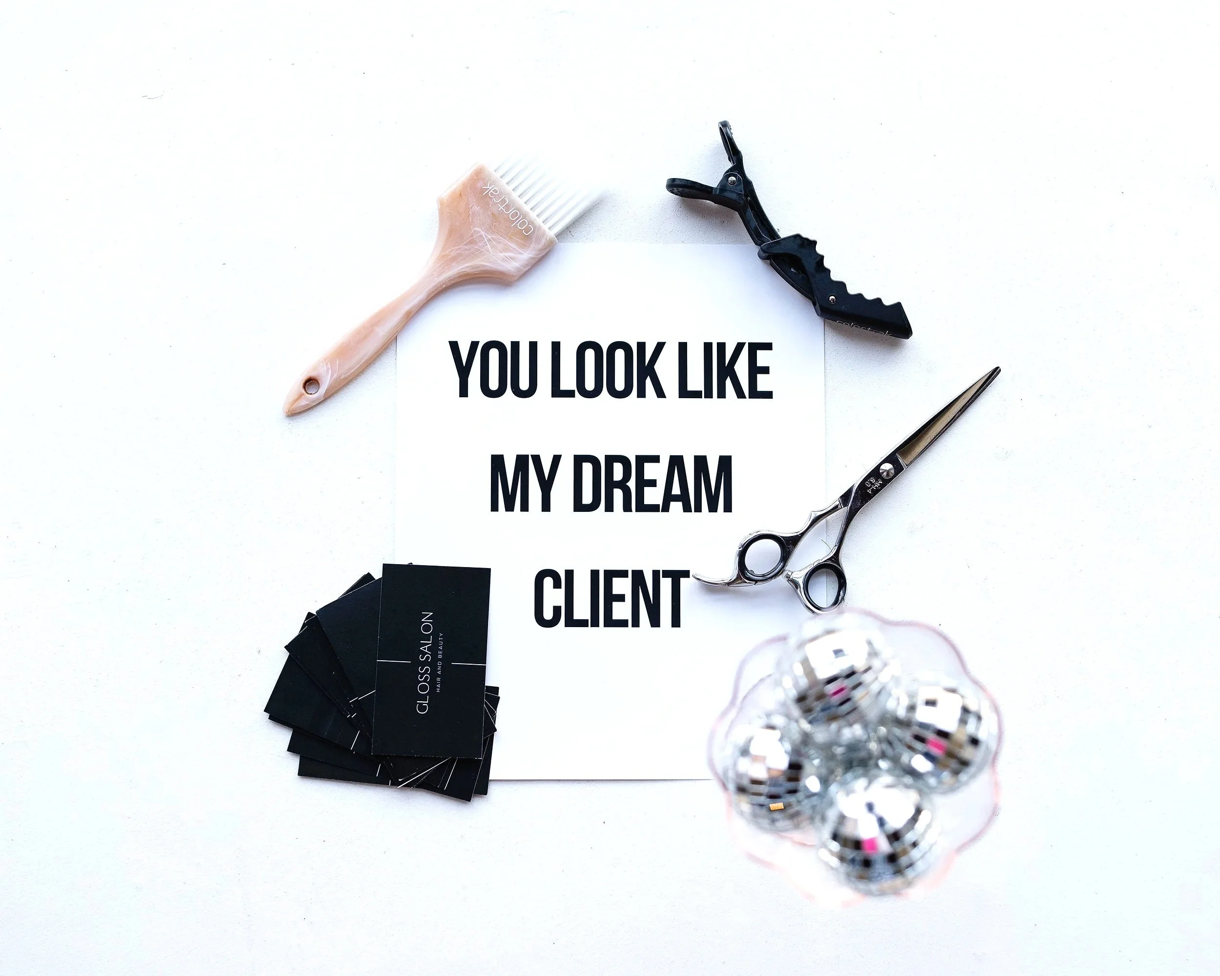 A flat lay of beauty and business items on a white surface, including scissors, a black hair color sample palette, a flared black clip, business cards, and a printed sign with the message "You look like my dream".