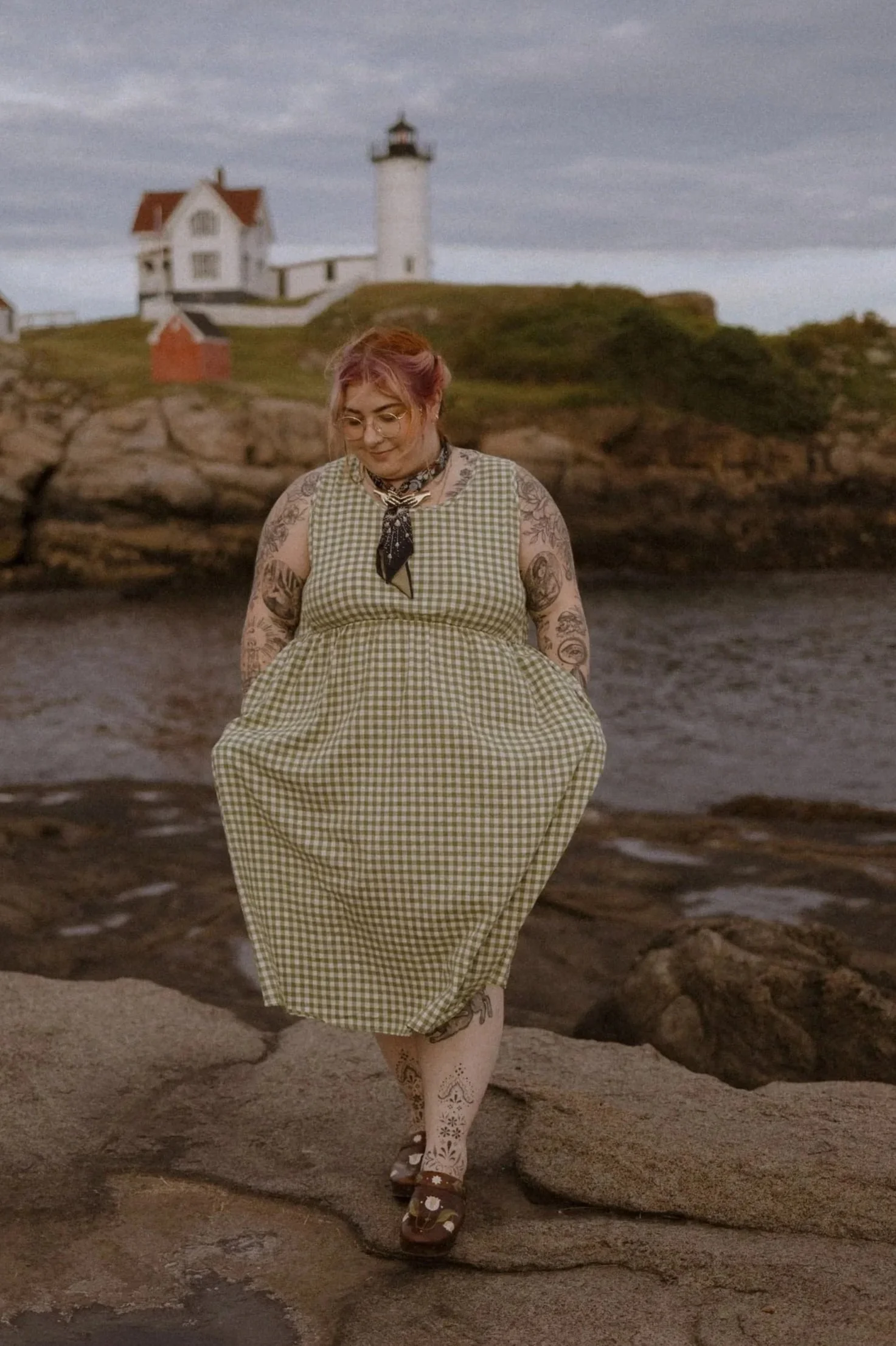 Alyssa stands in front a lighthouse in Maine wearing a green and white gingham dress and clogs they hand-painted themselves.