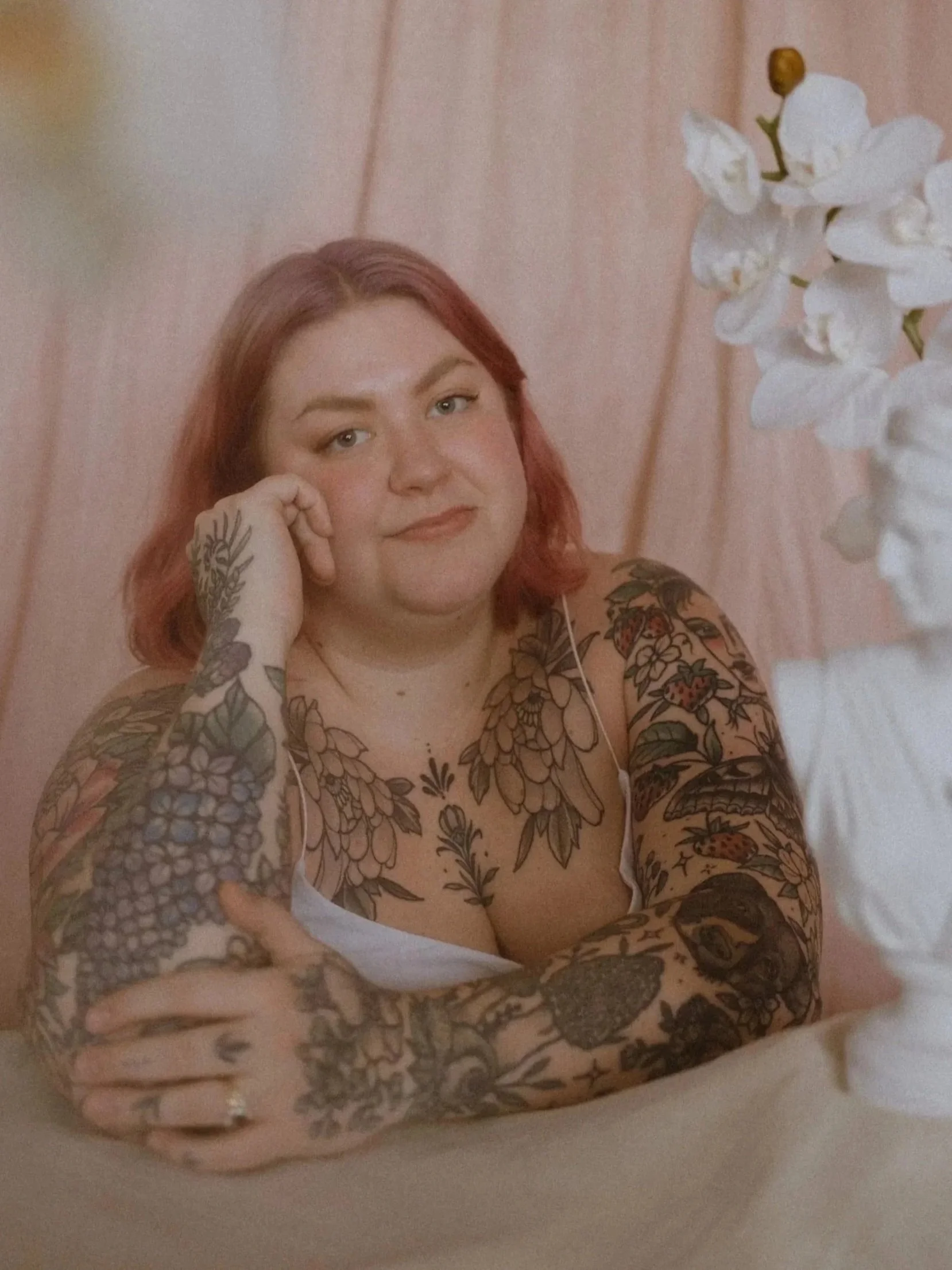 Holly leans on a table with her hand on her cheek, her other hand holds her elbow while she smiles at the camera. Her arms are tattooed with colorful flowers, fruit, and other nature motifs.