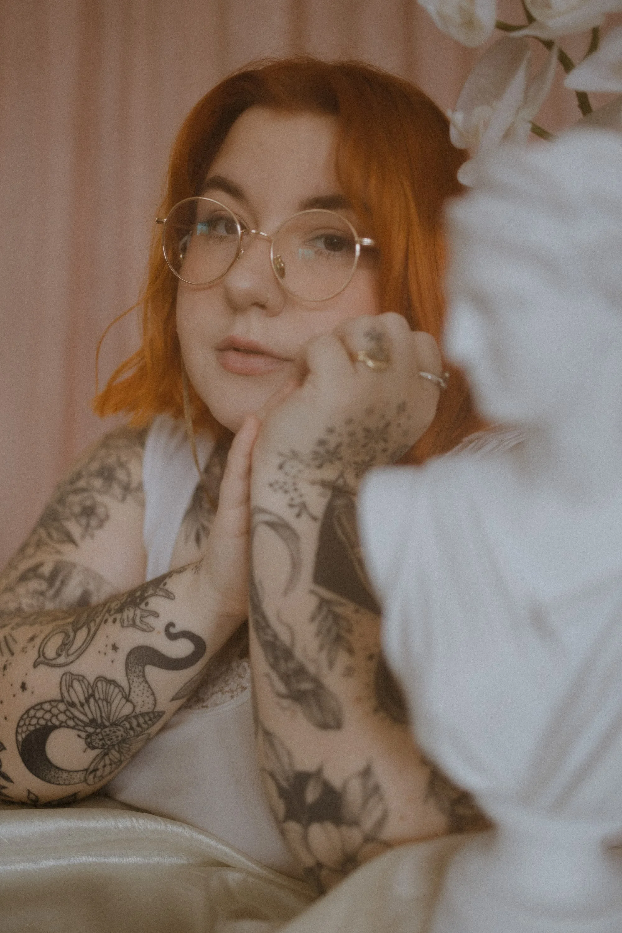 Alyssa poses with their hands folded next to their face. She's tattooed with various oddities and memories using only black ink. They have orange hair and gold rimmed circular glasses.