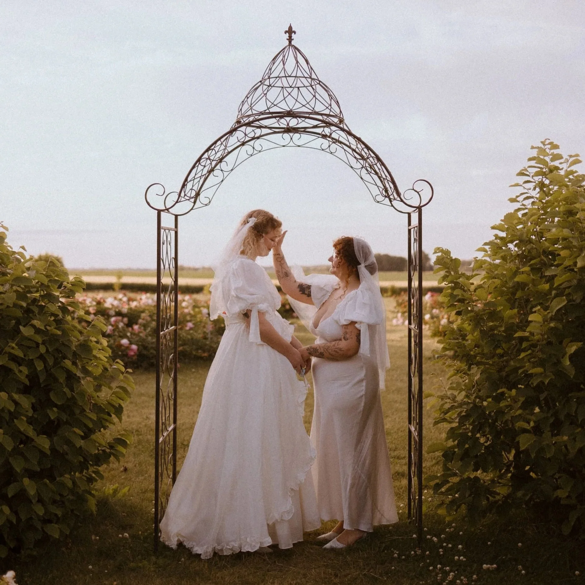 Queer couple stands under a decorative metal arch in a peony farm during their Love, The Underwoods elopement