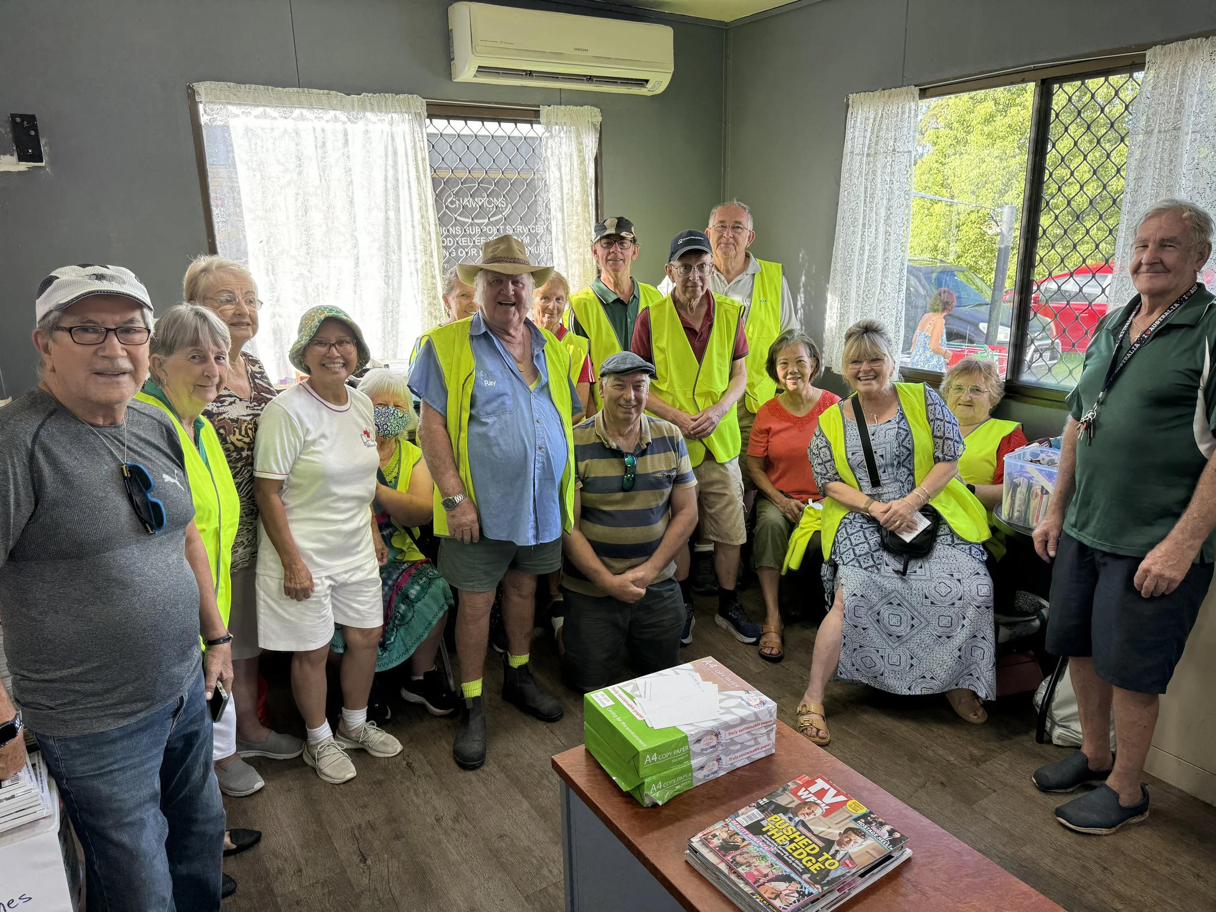 Group of people indoors, many wearing yellow reflective safety vests, smiling for the photo. The room has windows, lace curtains, and an air conditioning unit. Sitting on a table in the foreground are boxes of tissues and magazines.