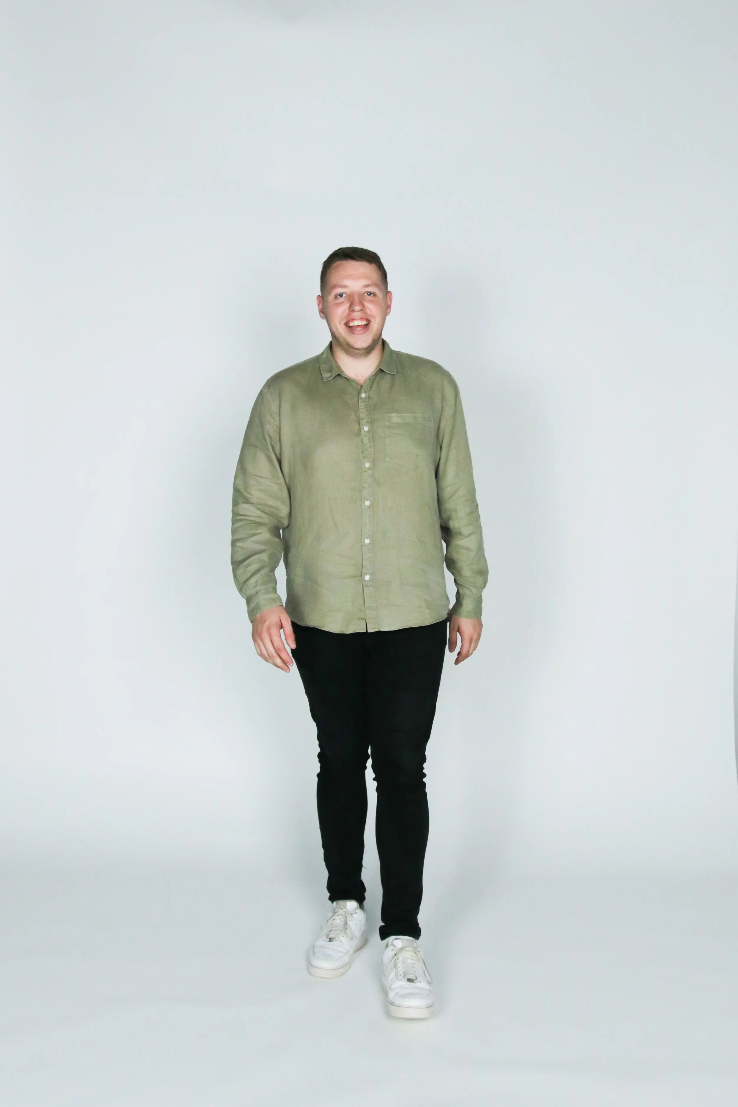 A young man with short brown hair, wearing a light green button-up shirt, black pants, and white sneakers, standing and smiling against a plain white background.