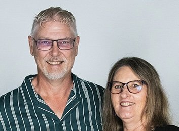 A smiling man and woman wearing glasses, standing together against a plain white background.