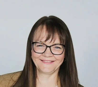 A woman with long brown hair, glasses, and a big smile, wearing a tan blazer against a light gray background.