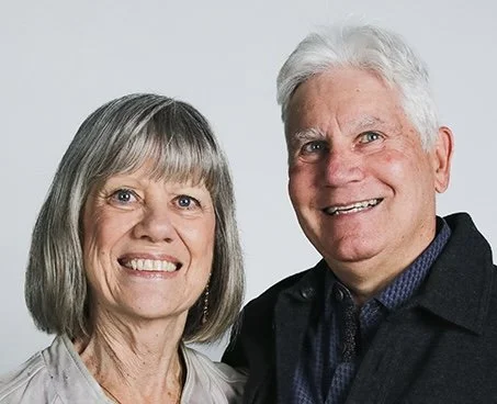 A smiling senior woman with gray hair and a senior man with gray hair, both wearing casual clothing, posing together against a light gray background.