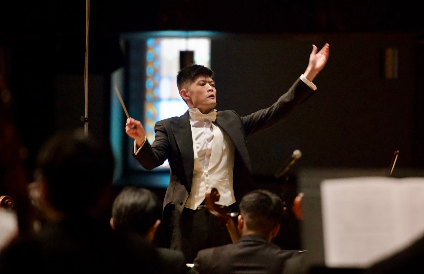 A male orchestra conductor in a tuxedo leading an orchestra indoors.