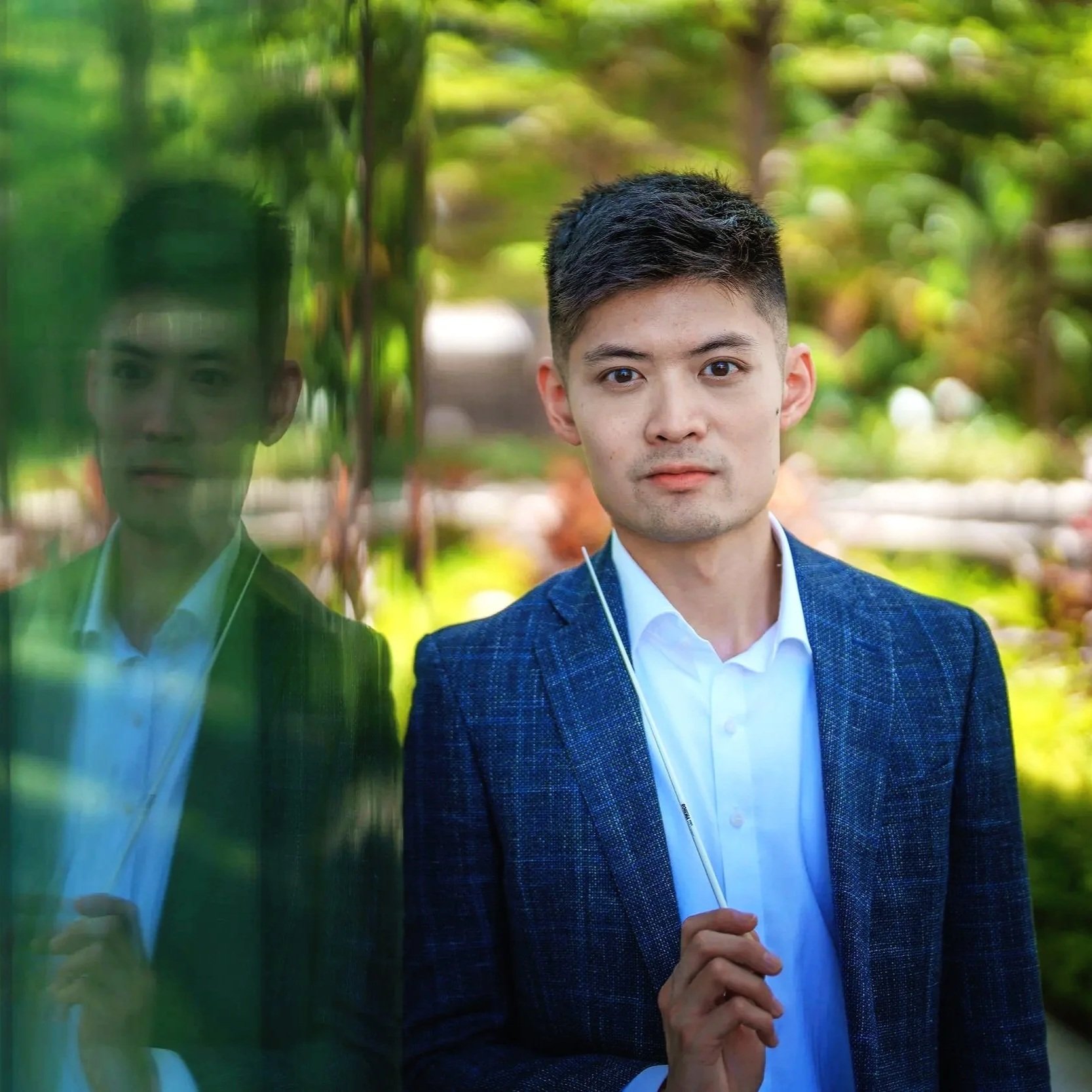 Young man with short black hair wearing a white shirt and blue blazer, holding a transparent umbrella with greenery in the background.