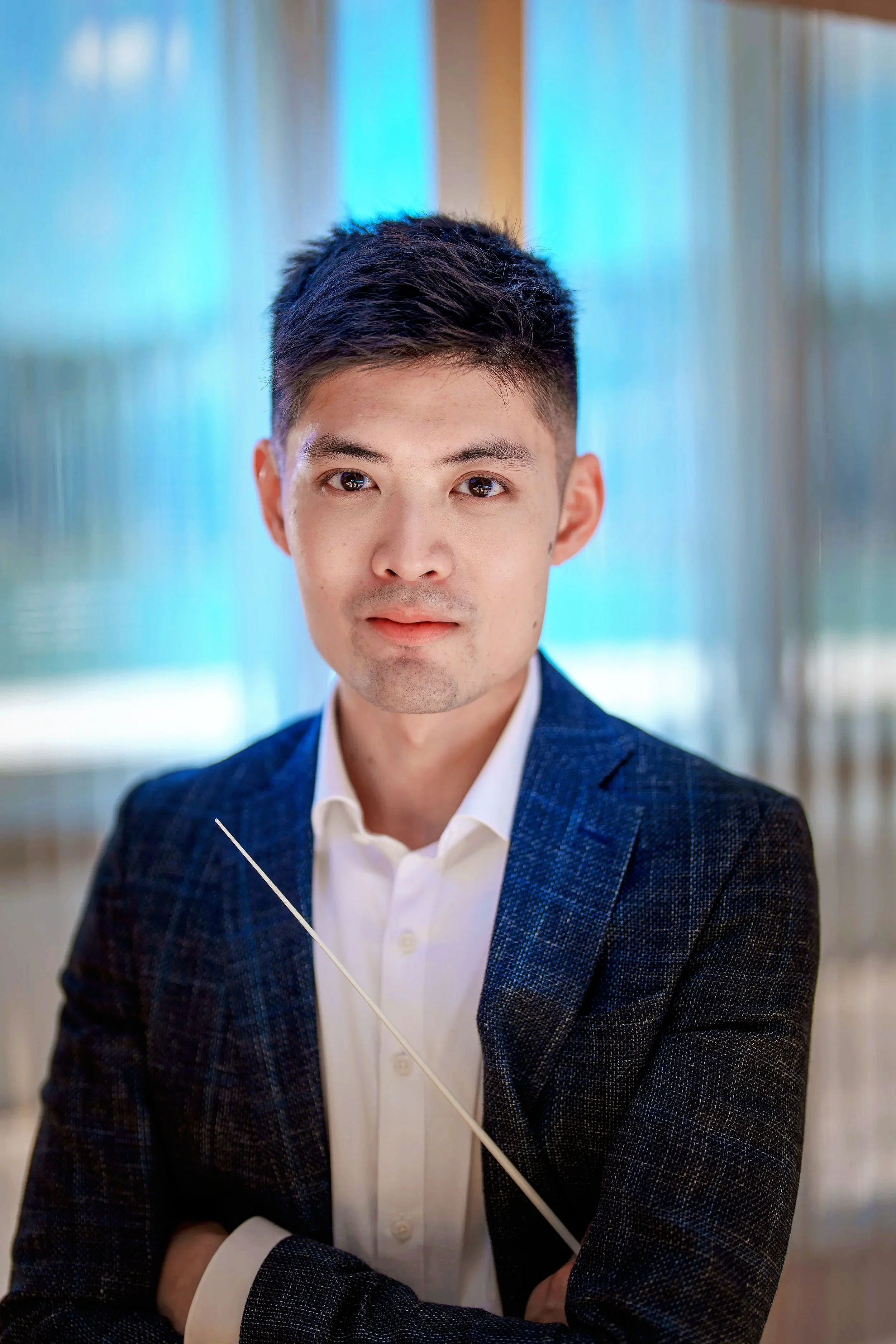 Headshot of a young Asian man in a white shirt and navy blazer, standing indoors with blurred background, looking directly at the camera.