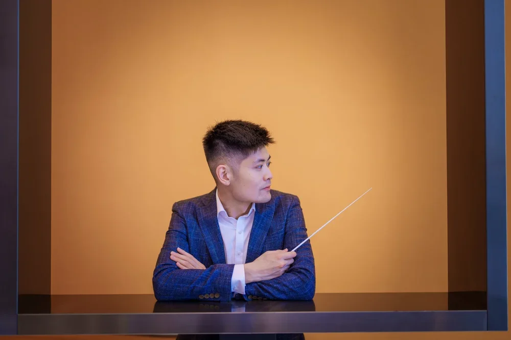 A man in a blue suit is sitting at a table with an orange background, holding a pointer stick and looking to his left.